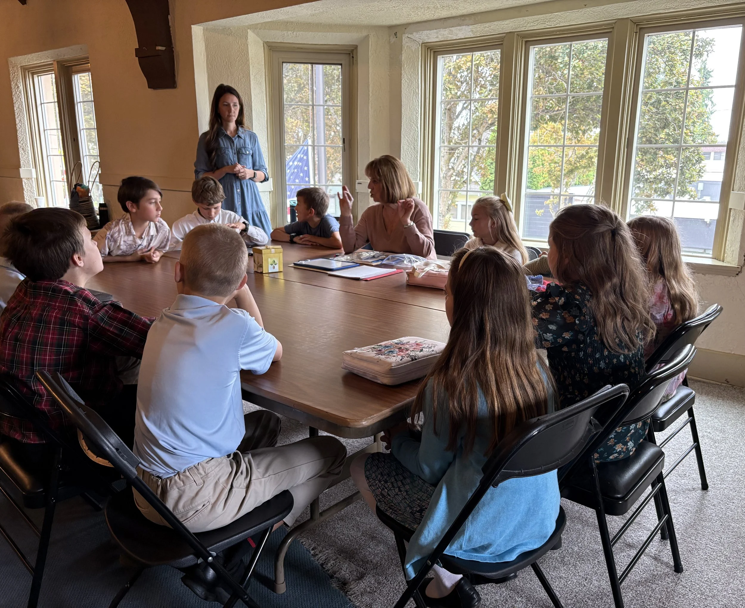 Children sitting around a large table in a bright room with large windows, listening to a woman speaking.
