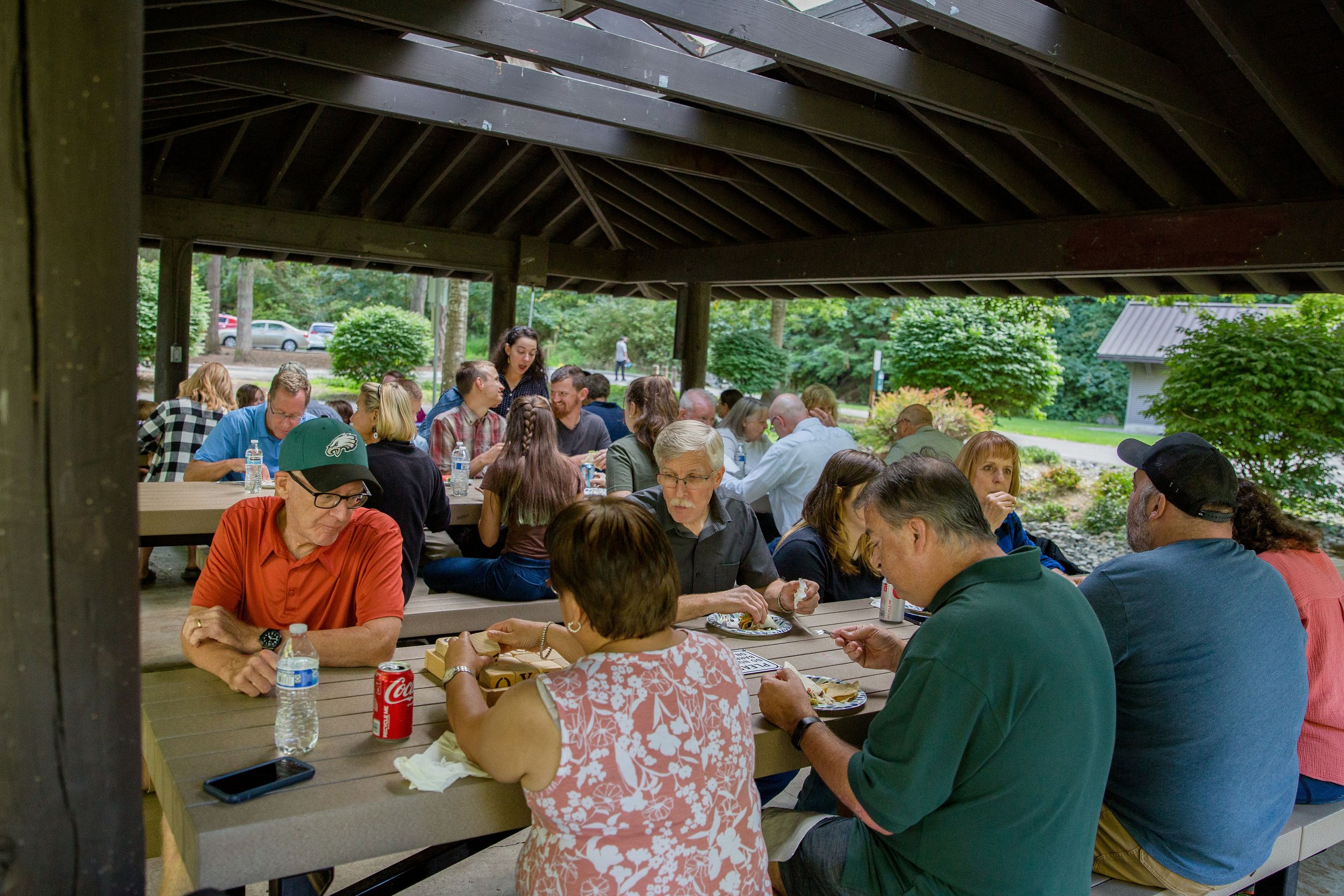 People gathered at picnic tables under a pavilion, eating and talking in an outdoor setting with trees and greenery in the background.