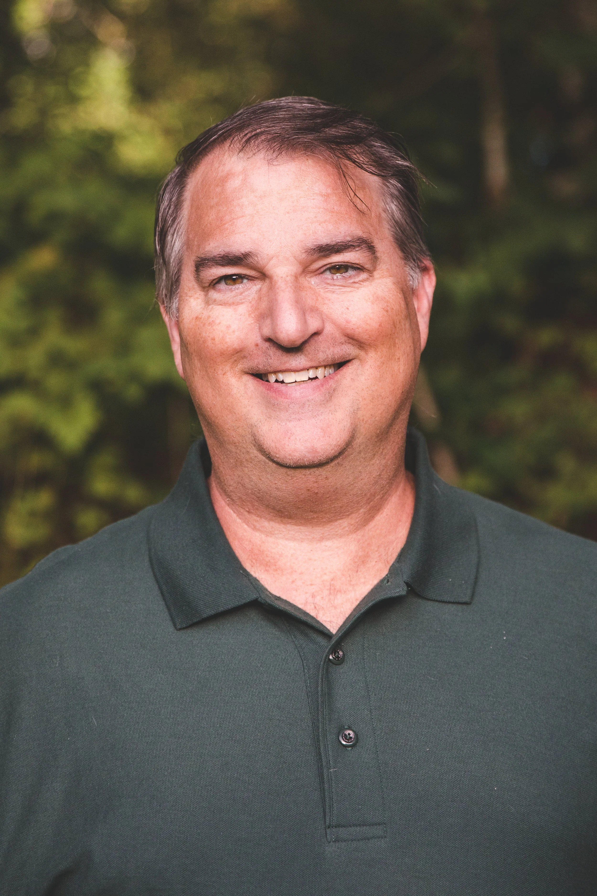 A smiling middle-aged man with short dark hair wearing a dark green polo shirt, standing outdoors with a blurred green foliage background.