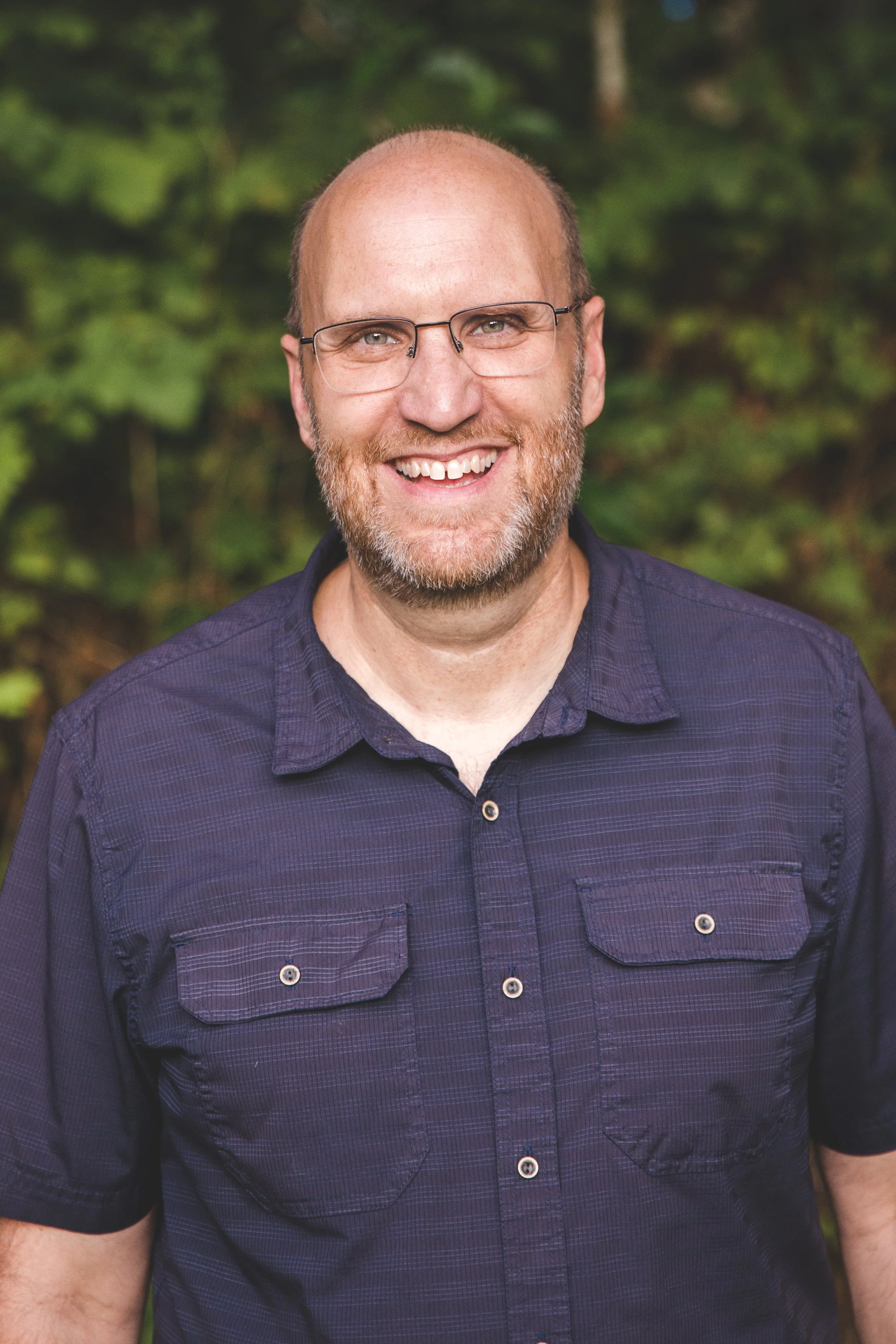 A smiling man with glasses and a beard, wearing a dark button-up shirt, standing outdoors with green foliage in the background.