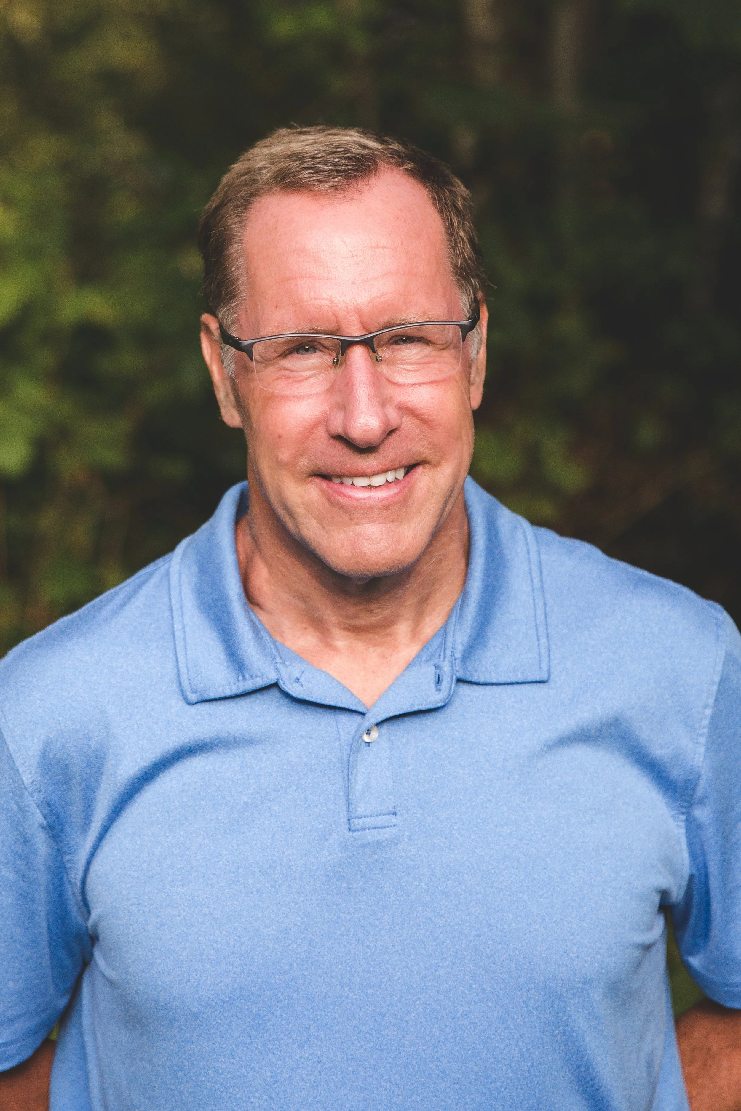 A man with glasses and short brown hair smiling, wearing a blue polo shirt, outdoors with a green forest background.