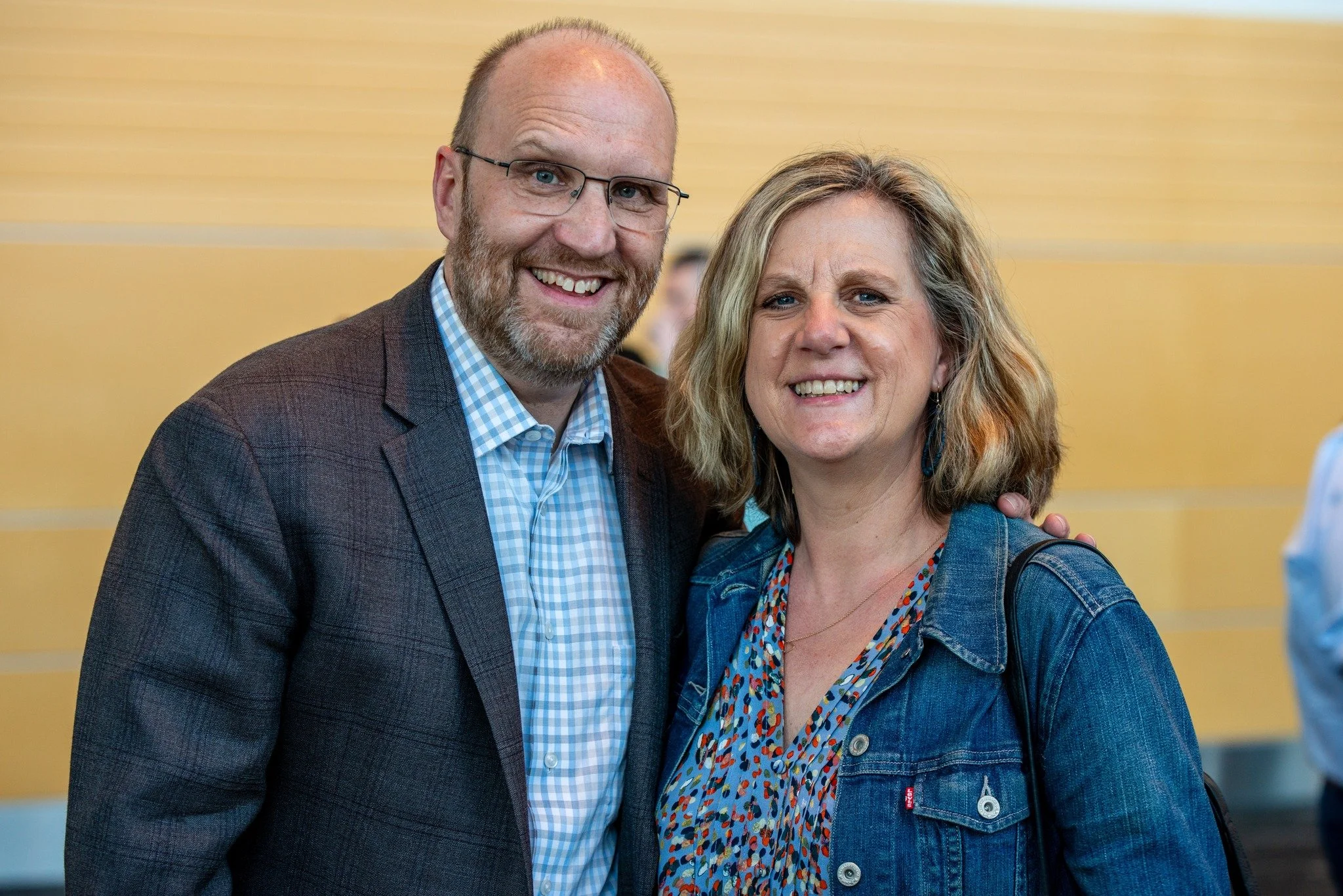 The senior pastor and his wife smiling and posing together at an indoor event. The man has a beard, glasses, and is wearing a plaid shirt and blazer. The woman has shoulder-length wavy hair and is wearing a denim jacket over a colorful blouse.