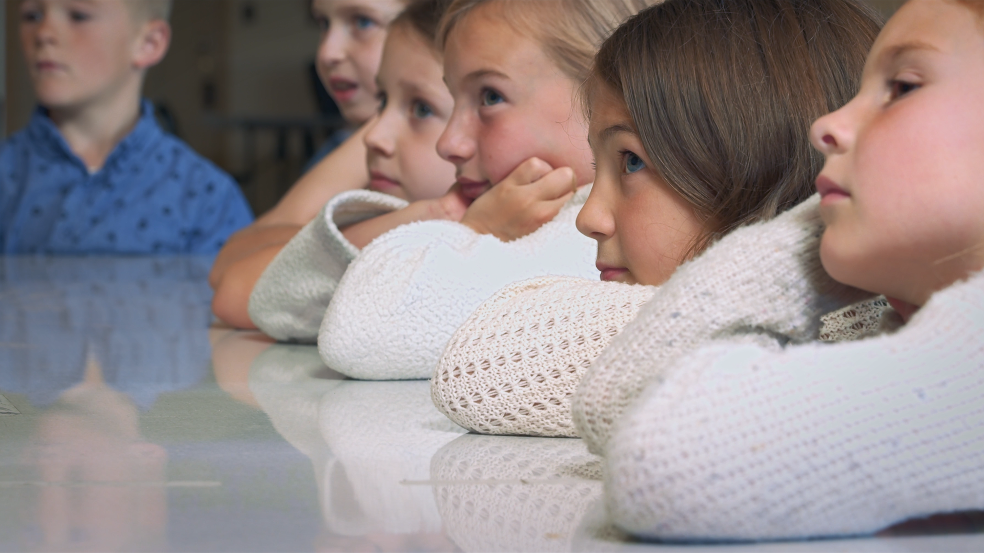 Group of young children sitting at a table, resting their chins on folded arms, attentively listening.