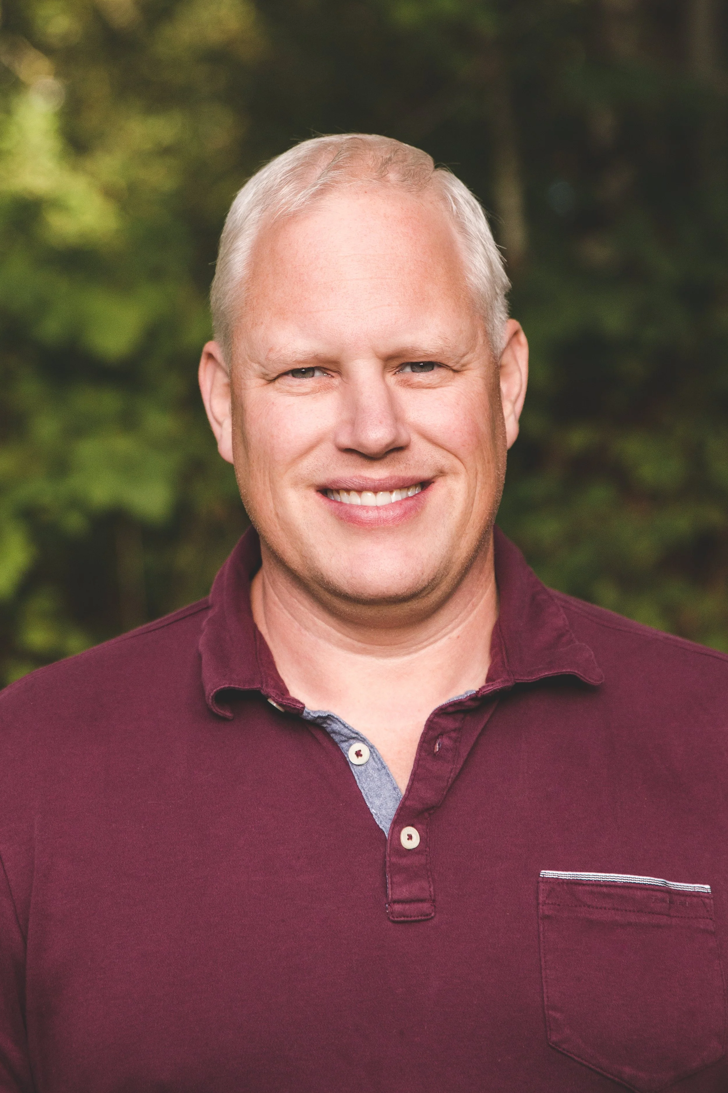 Headshot of a smiling man with short, light-colored hair wearing a maroon shirt, outdoors with a blurred green foliage background.