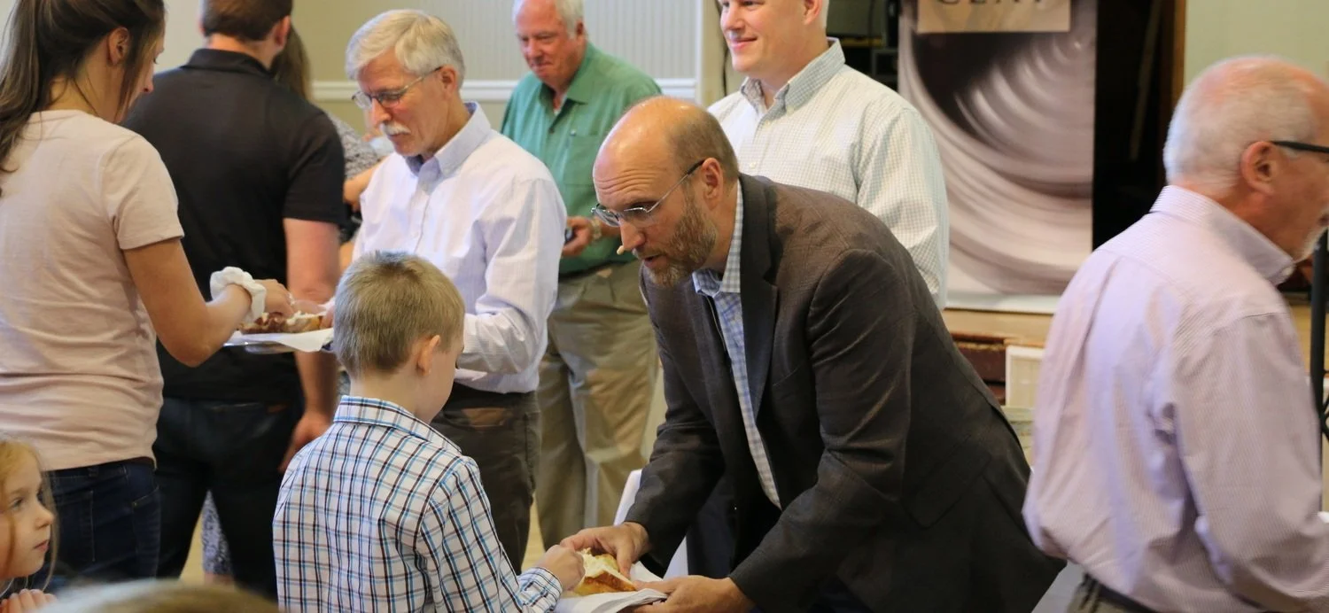 The senior pastor bends down to offer communion bread to a young boy wearing a plaid shirt at a Sunday service with several other people around.