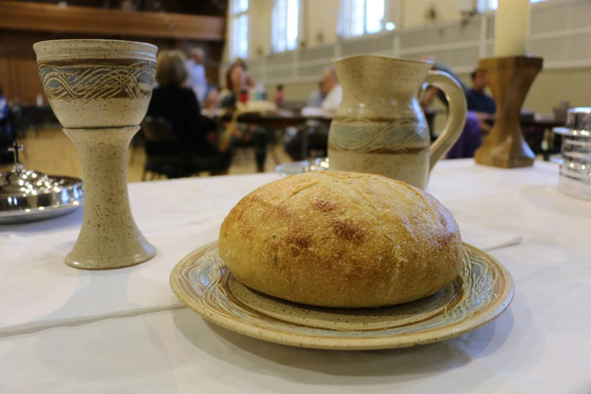 A loaf of bread on a decorative plate on a table set for communion, with ceramic goblets and pitchers in the background, and people sitting at tables in a dining hall.