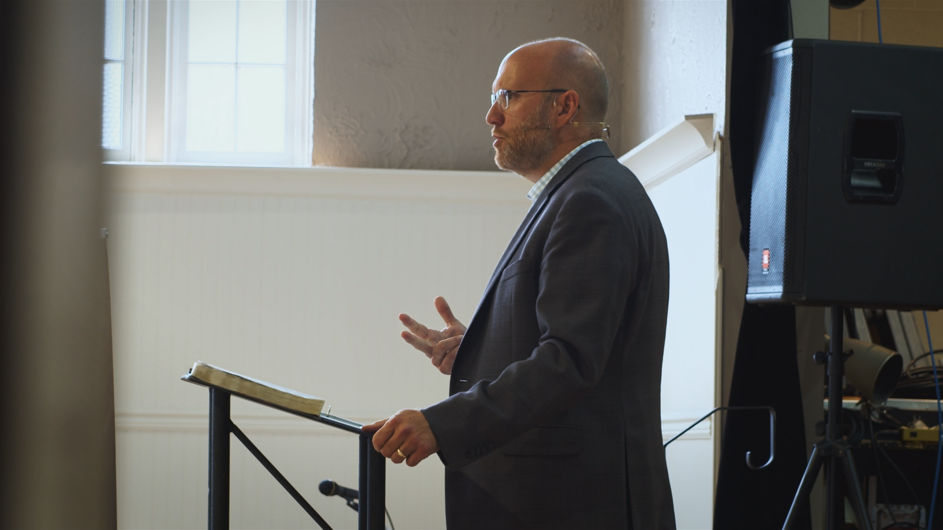 The senior pastor in a suit giving a sermon, standing at a podium with a book and gesturing with his hand, indoor setting with a window and speaker in the background.