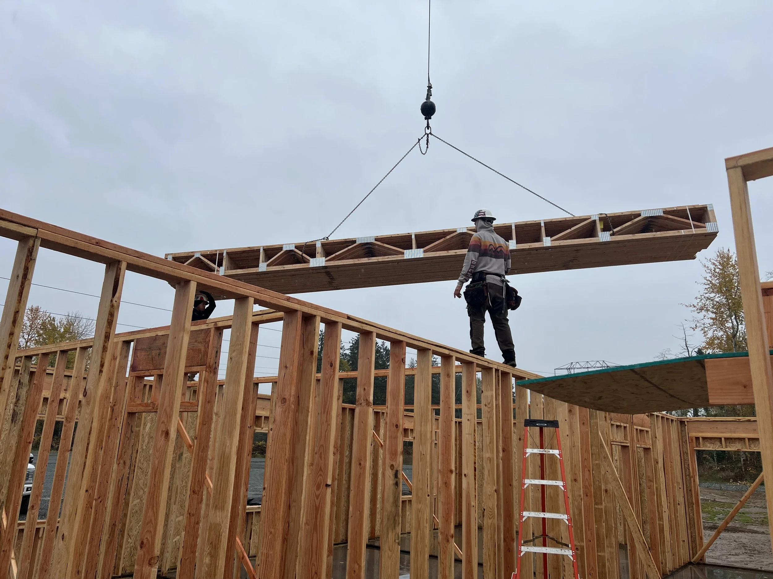 Construction workers building a wooden structure, with one worker walking on a platform and another on an upper level, while a crane lifts a large wooden beam overhead.