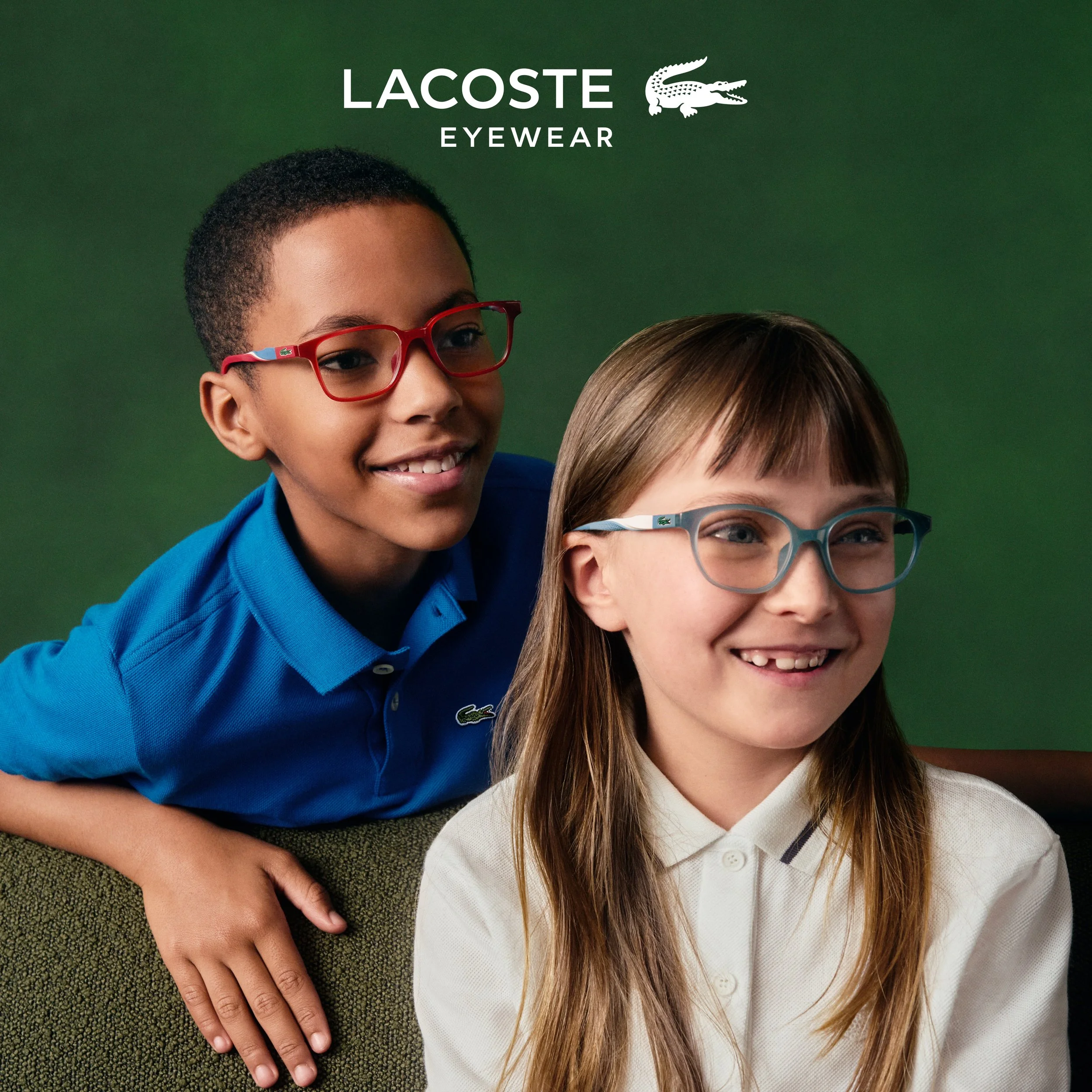 Two children, one with short curly hair and red glasses wearing a blue Lacoste polo shirt, and the other with long straight hair and blue glasses wearing a white shirt, smiling and sitting against a green background with Lacoste Eyewear branding.
