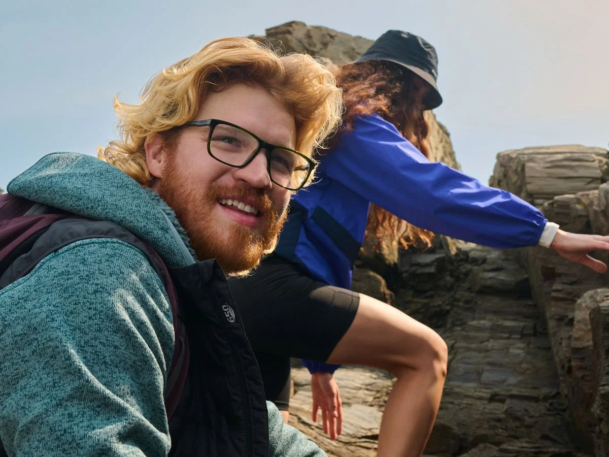 A man wearing glasses and a woman in a blue jacket rock climbing outdoors on a rocky surface.