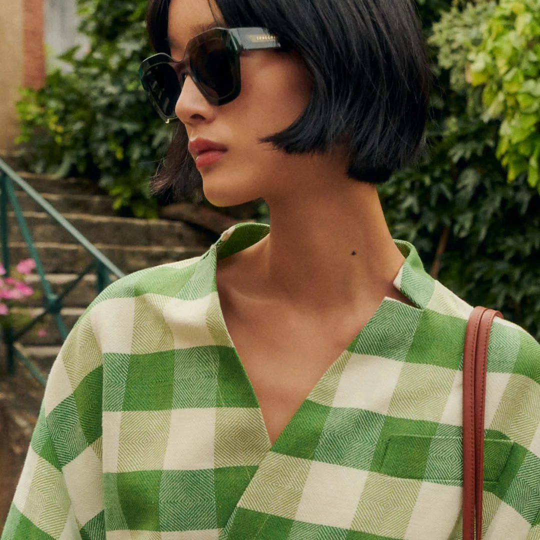 A woman with short black hair wearing large dark sunglasses and a green and white checkered shirt, standing outdoors with green foliage and staircase in the background.