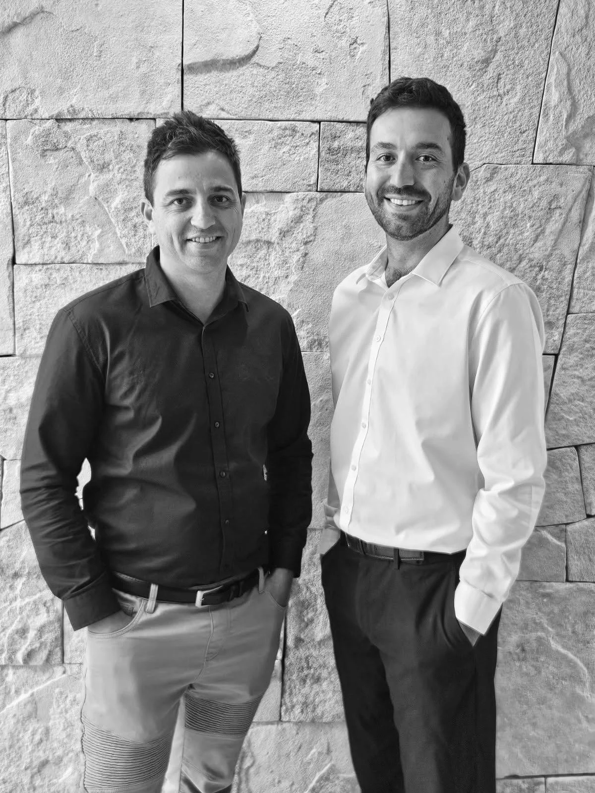 Two men standing side by side in front of a stone wall, smiling at the camera.