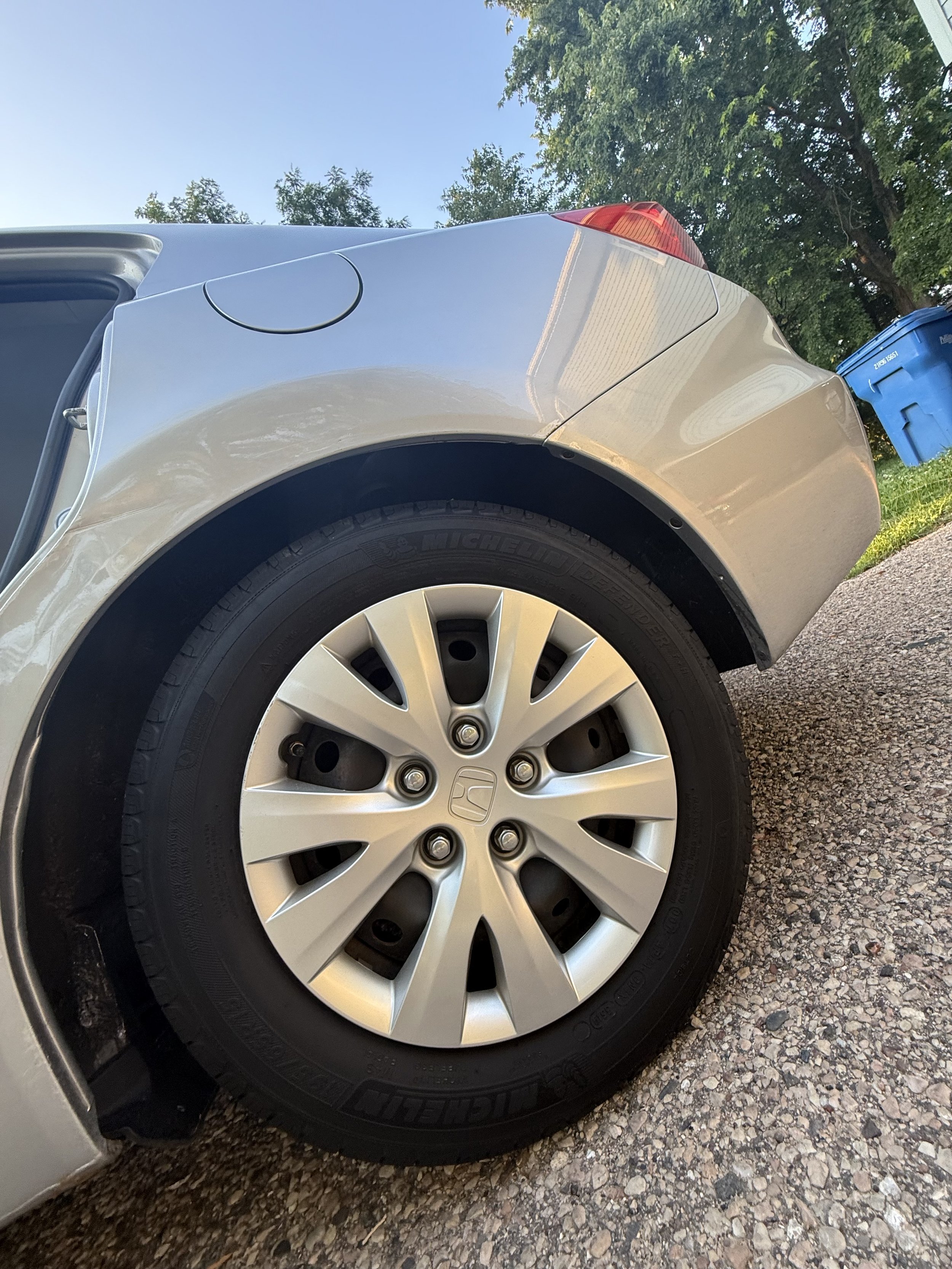 Close-up of a silver Honda car's rear wheel and tire parked on a gravel driveway, with a blue recycling bin, green trees, and a blue sky in the background.