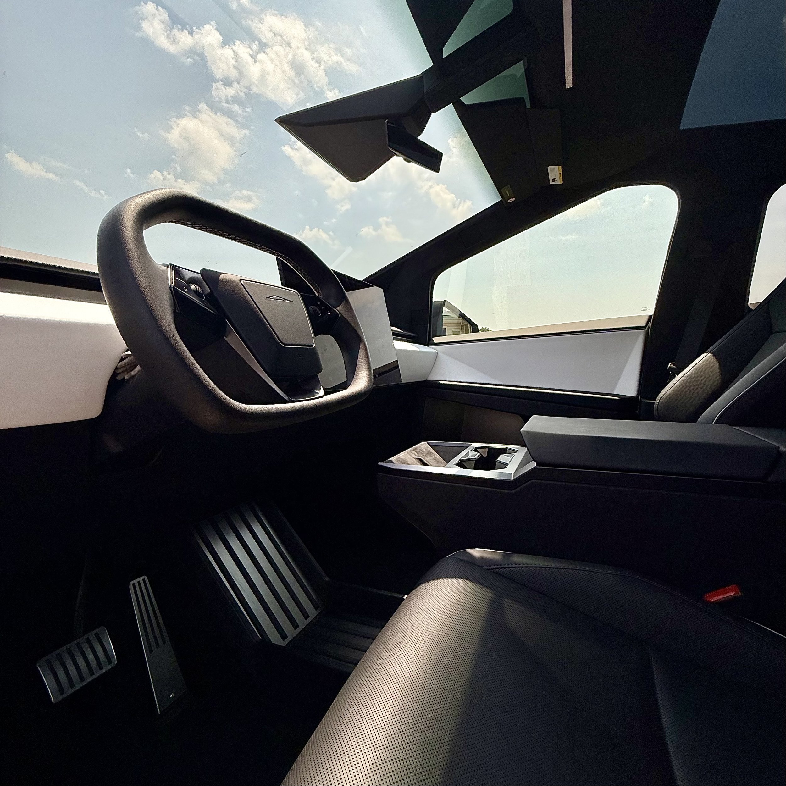 Interior of a futuristic electric vehicle with a minimalist dashboard, black steering wheel, digital display, and large panoramic windows showing a blue sky with clouds.