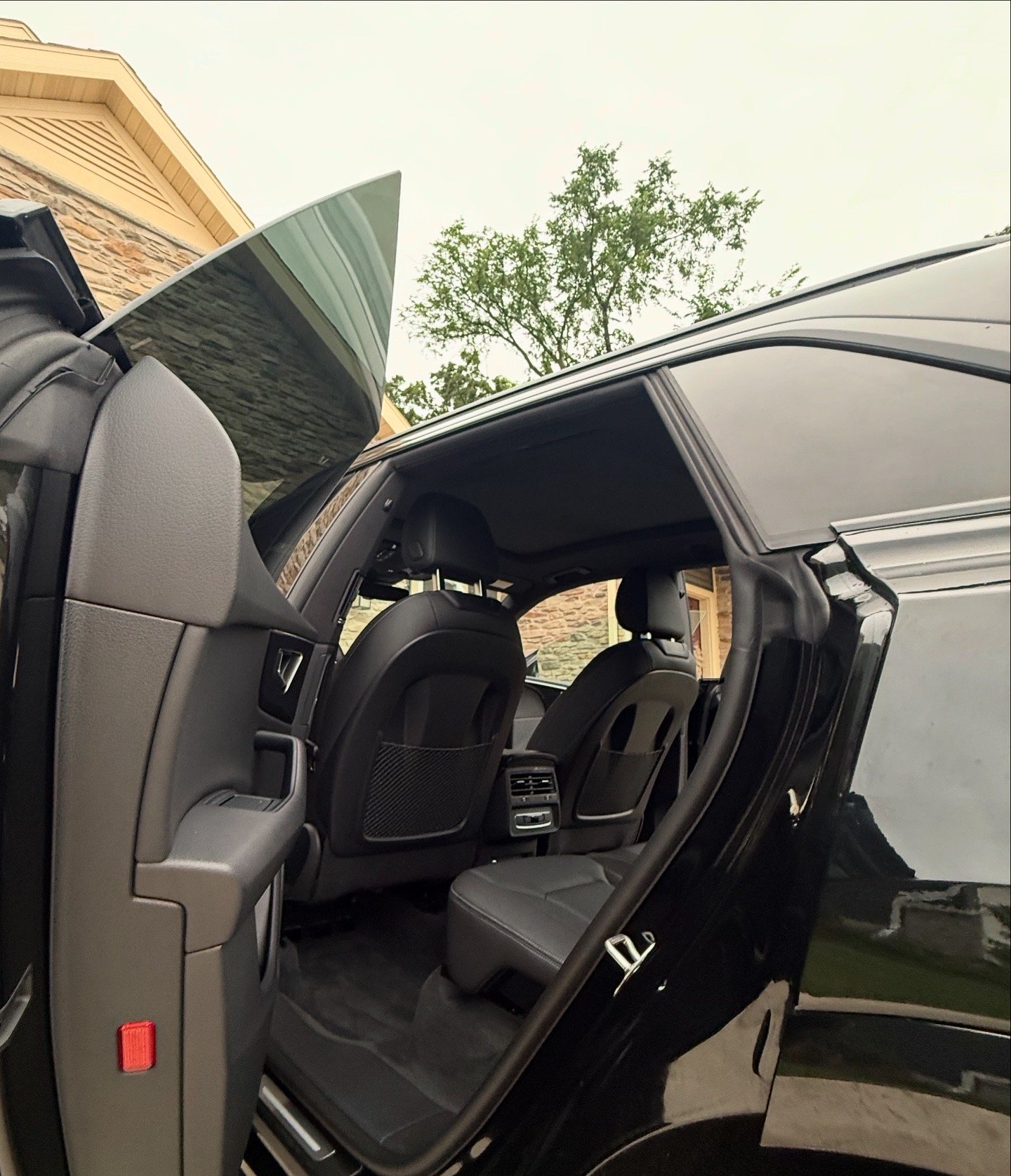 Inside view of a black SUV with the open rear passenger door, showing black leather seats, rear air vents, and the back of the front seats, with a background of trees and houses.