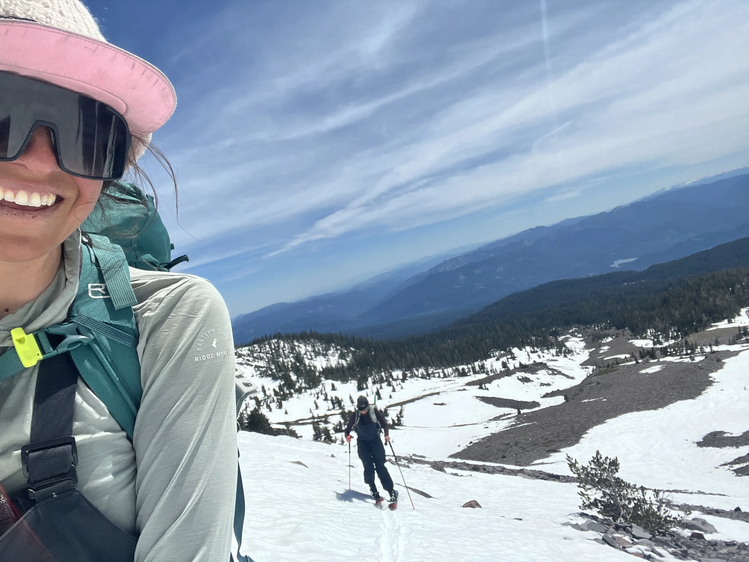 A woman taking a selfie while snowshoeing in a snow-covered mountainous area with blue skies, with others snowshoeing in the background.