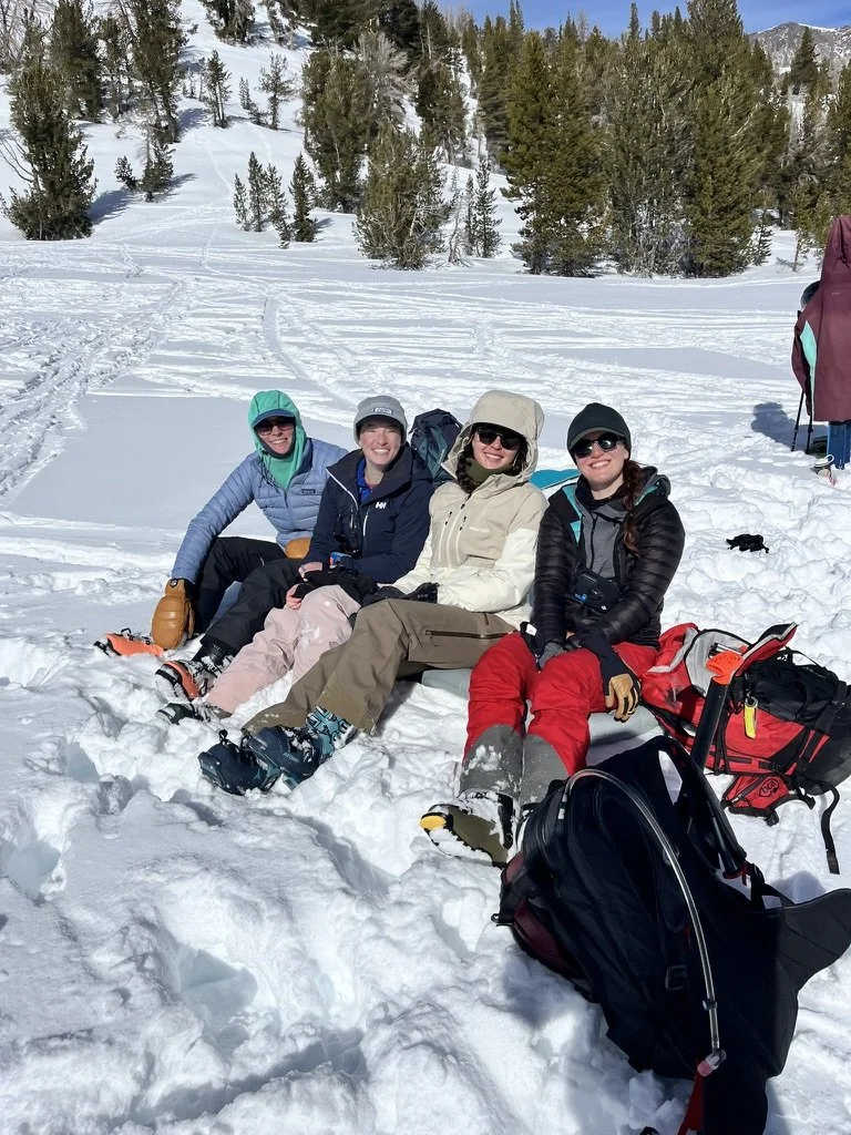 Four women sitting on snow in a winter landscape with trees and mountains in the background, dressed in winter gear and surrounded by backpacks and gear.