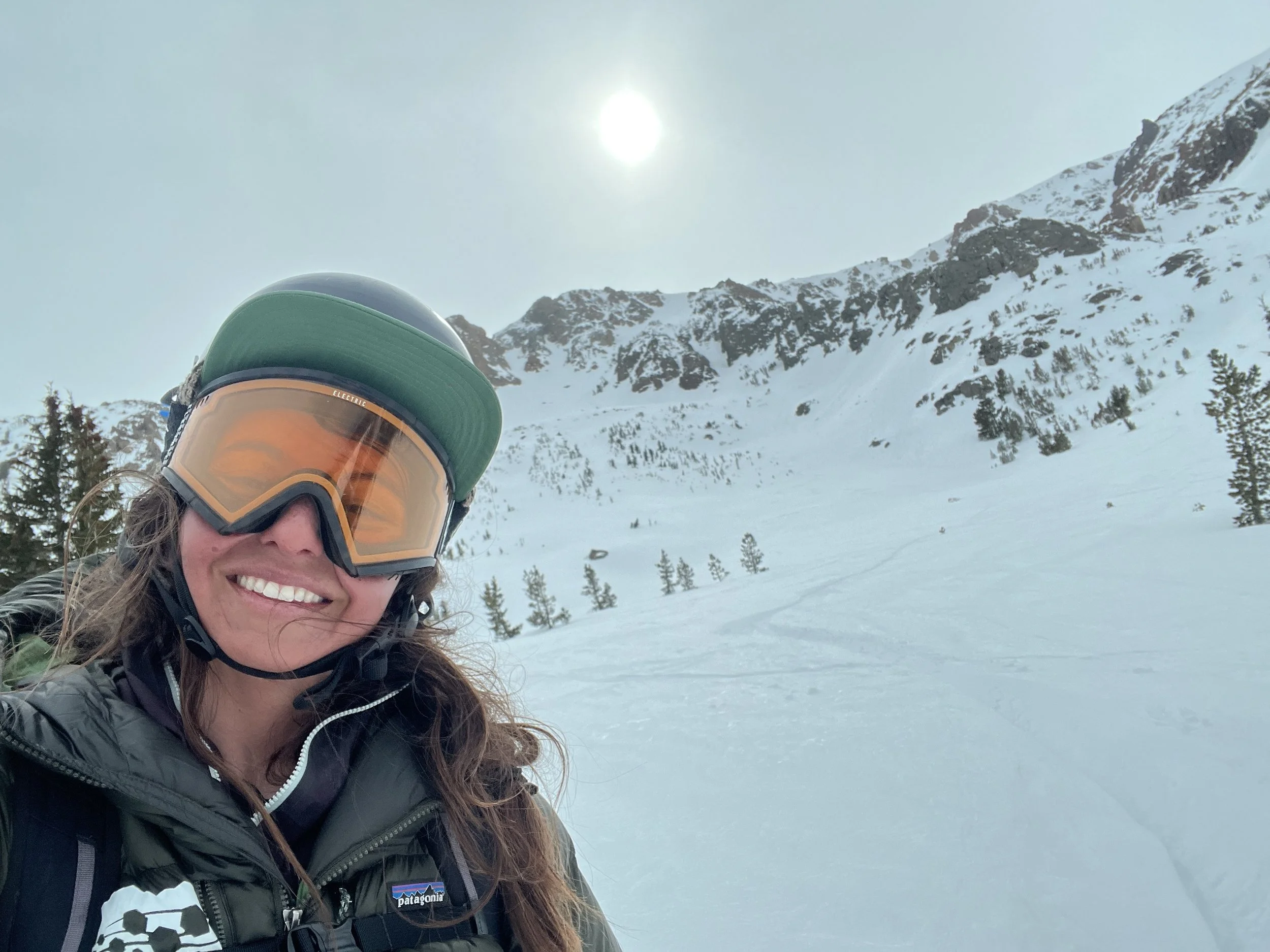 A woman in ski gear, including a helmet and goggles, smiling in a snowy mountain landscape with trees and mountain peaks under a hazy sun.