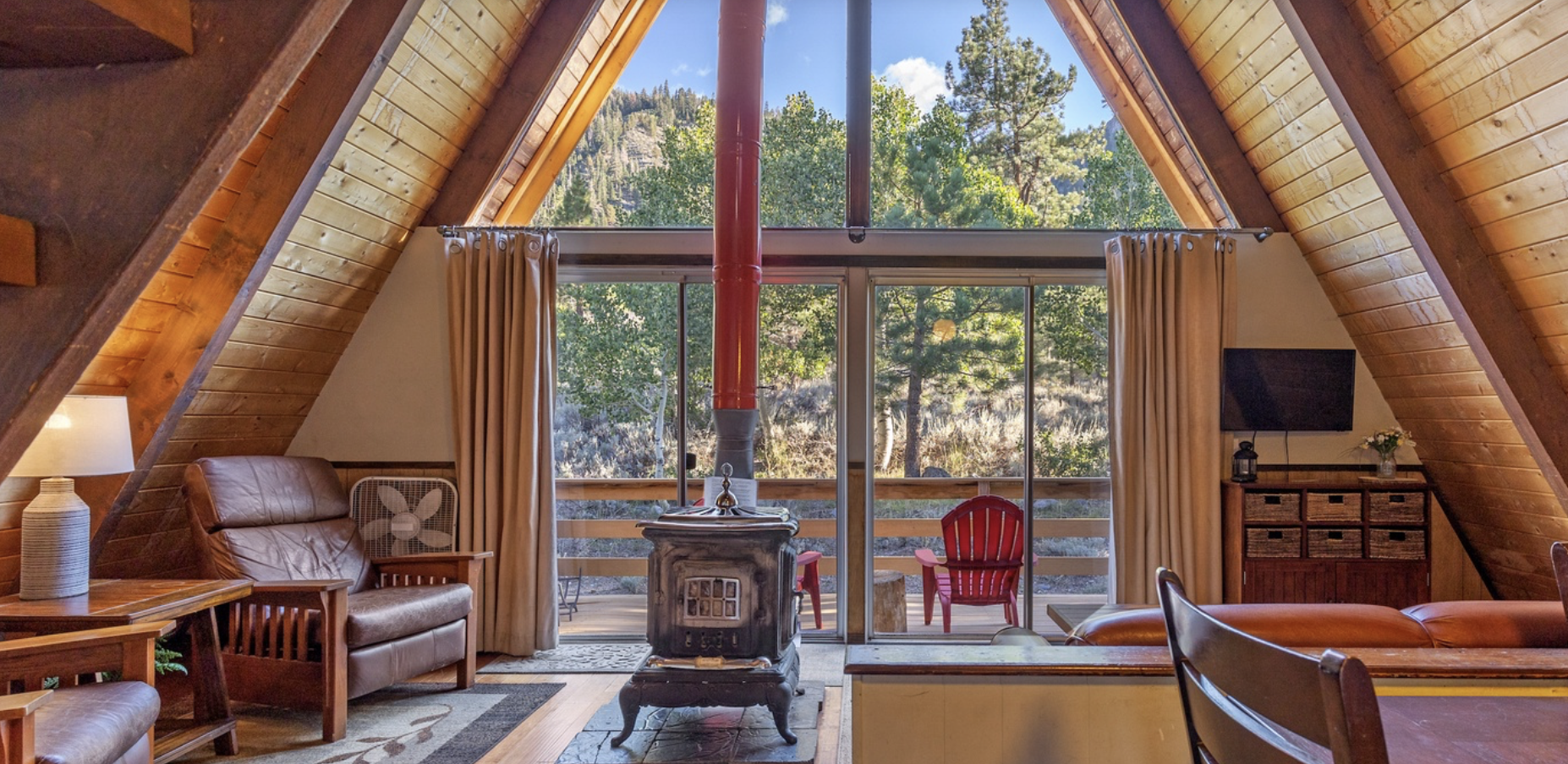 Interior of a cozy A-frame cabin living room with vaulted wooden ceiling, a stone fireplace with a red chimney pipe, large windows showing trees and mountains, leather furniture, a wooden cabinet, and a ceiling fan.