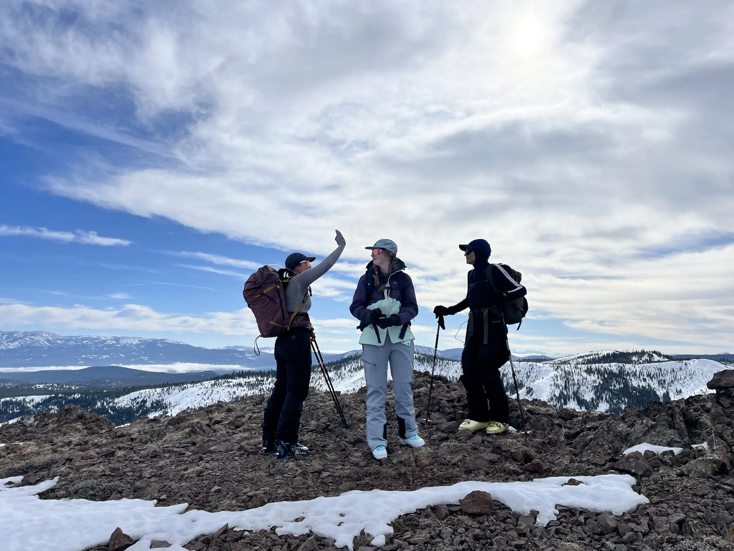 Four hikers standing on rocky terrain in a snowy mountainous landscape, with snow-capped peaks in the background and a partly cloudy sky.