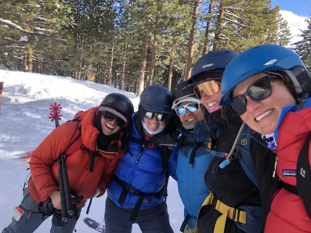 Group of five people in winter gear taking a selfie on snowy terrain surrounded by tall pine trees, smiling and wearing helmets and sunglasses.
