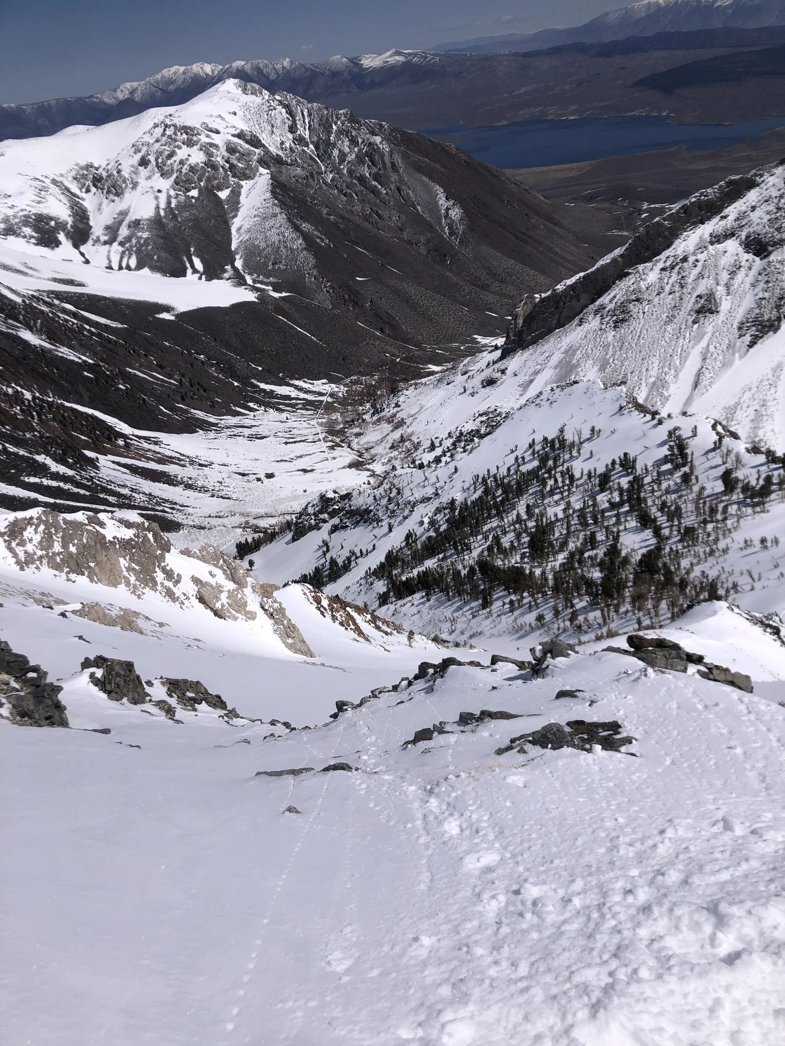 Snow-covered mountain trail descending into a valley with sparse trees, rugged rocky slopes, and a distant lake.