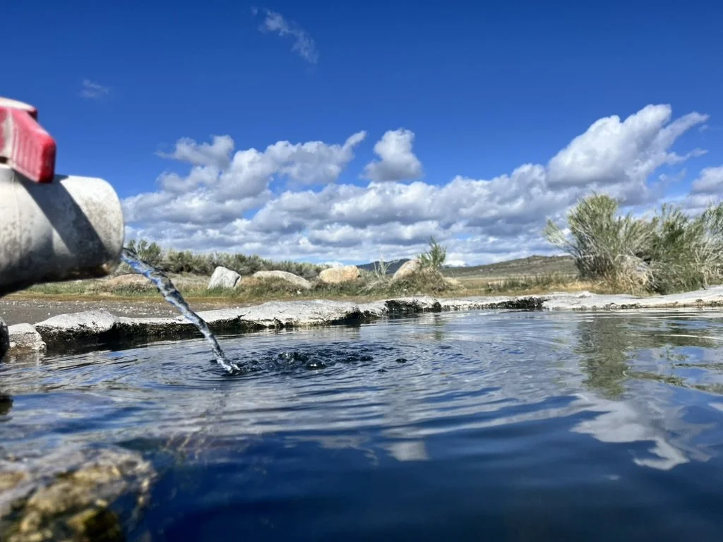 A close-up of a water source with a pipe on the left side releasing water into a natural stream, with grassy hills and a partly cloudy sky in the background.
