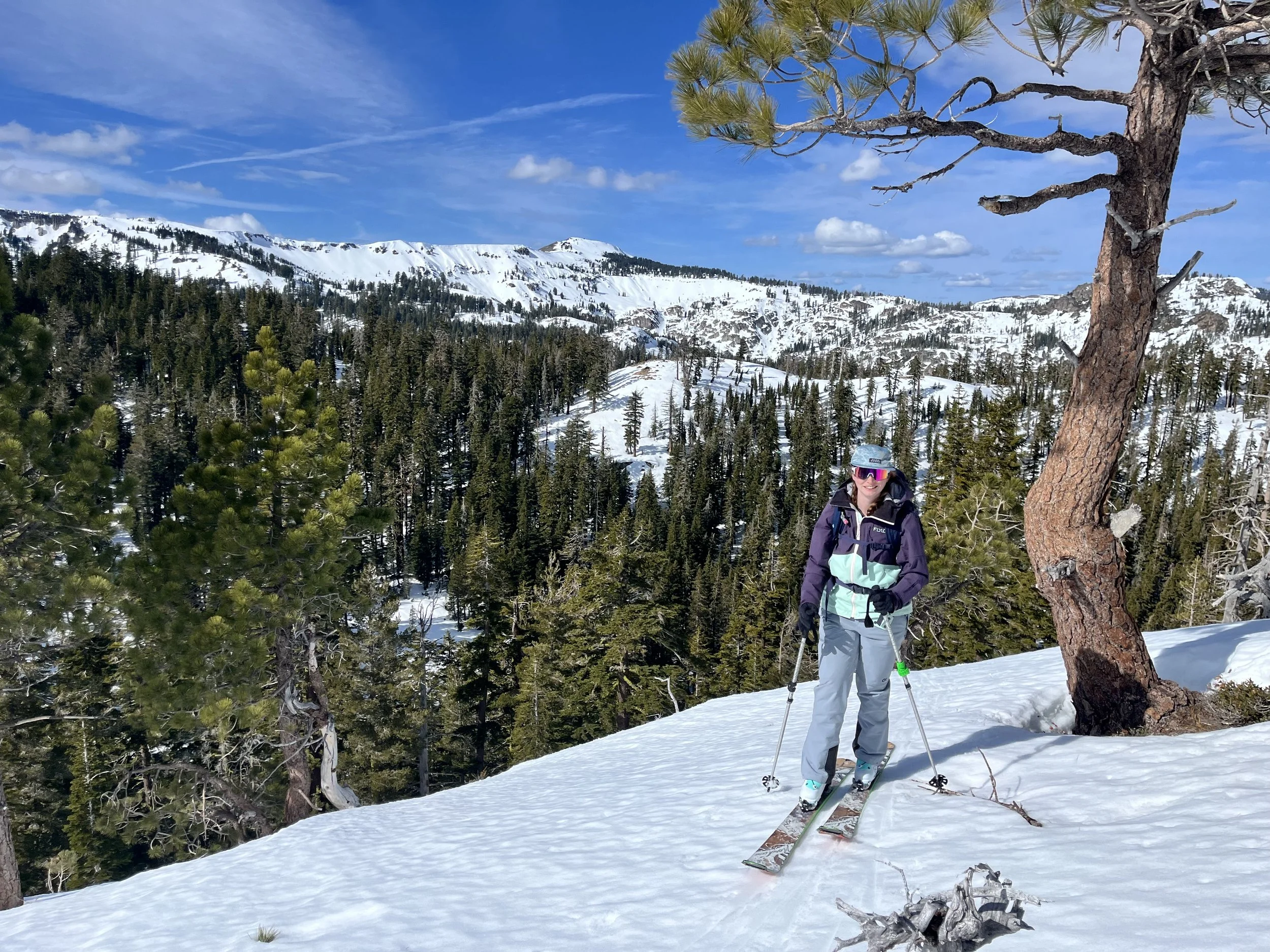 A person wearing ski gear and sunglasses standing on snow with ski poles in a mountainous, snowy landscape. There are trees and mountains in the background under a bright blue sky with scattered clouds.