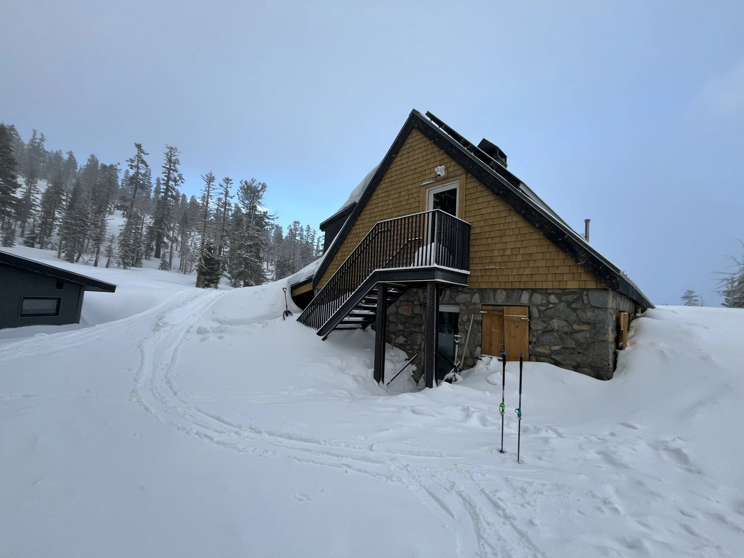 A snow-covered mountain house with a wooden exterior, stone foundation, black roof, and outdoor staircase, surrounded by snow and trees under a cloudy sky.