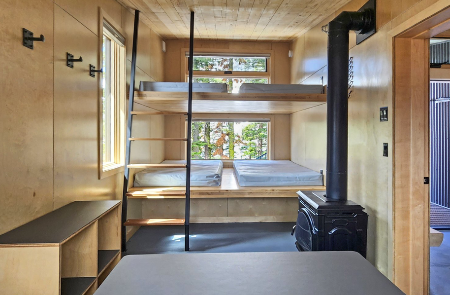 Interior of a small wooden cabin bedroom with a bunk bed, a wood stove, and minimal furniture.