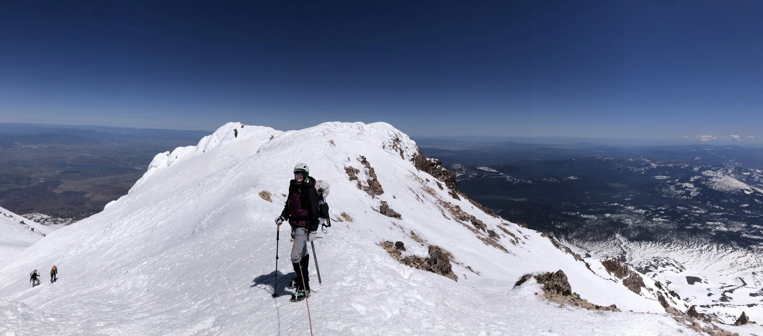 A person in winter gear standing on a snow-covered mountain summit with a snow-capped ridge and a distant valley below under a clear blue sky.