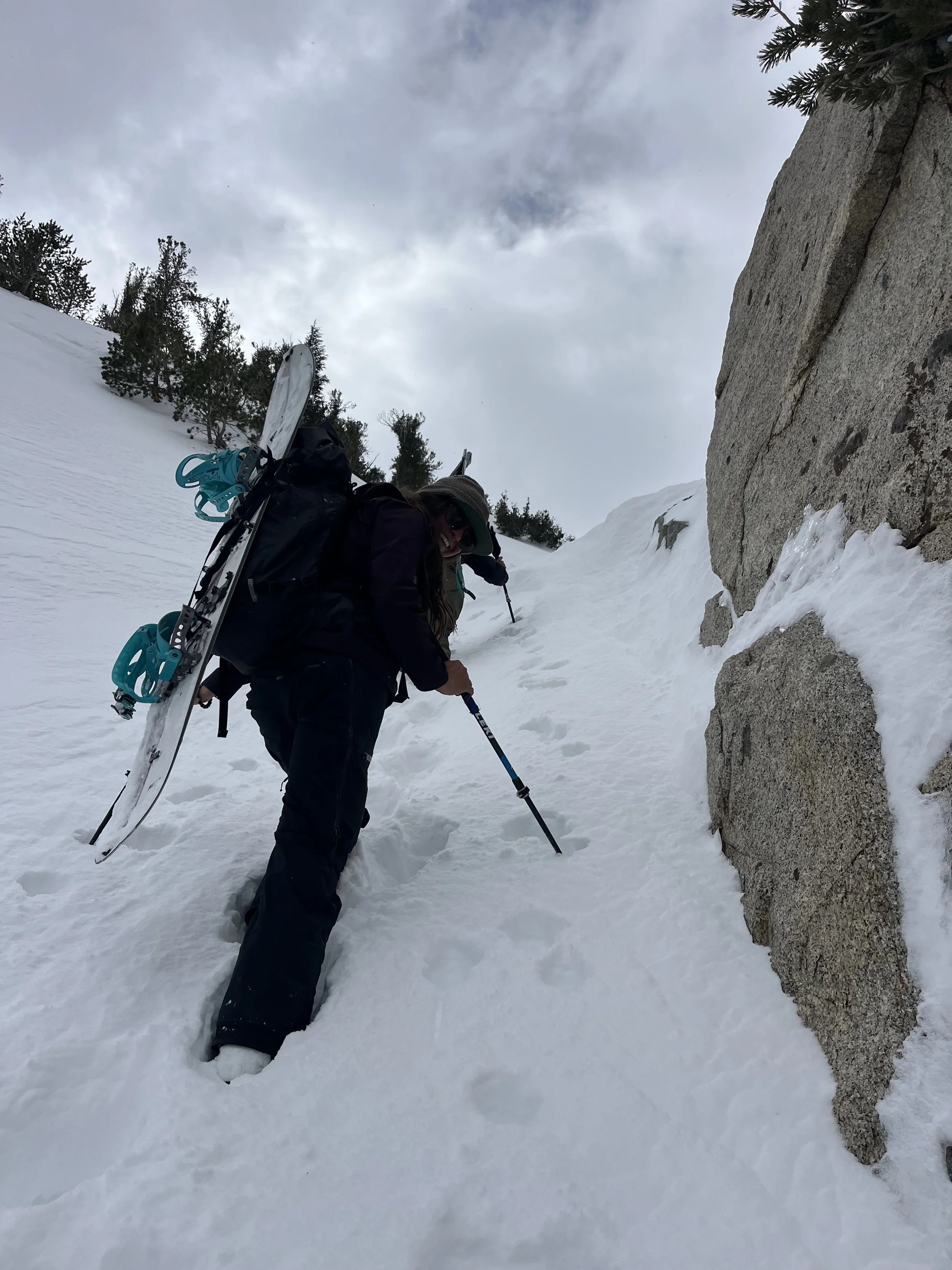 Person climbing a snow-covered mountain with a backpack, snow shoes, and trekking poles, next to a large rock and sparse trees in the background.