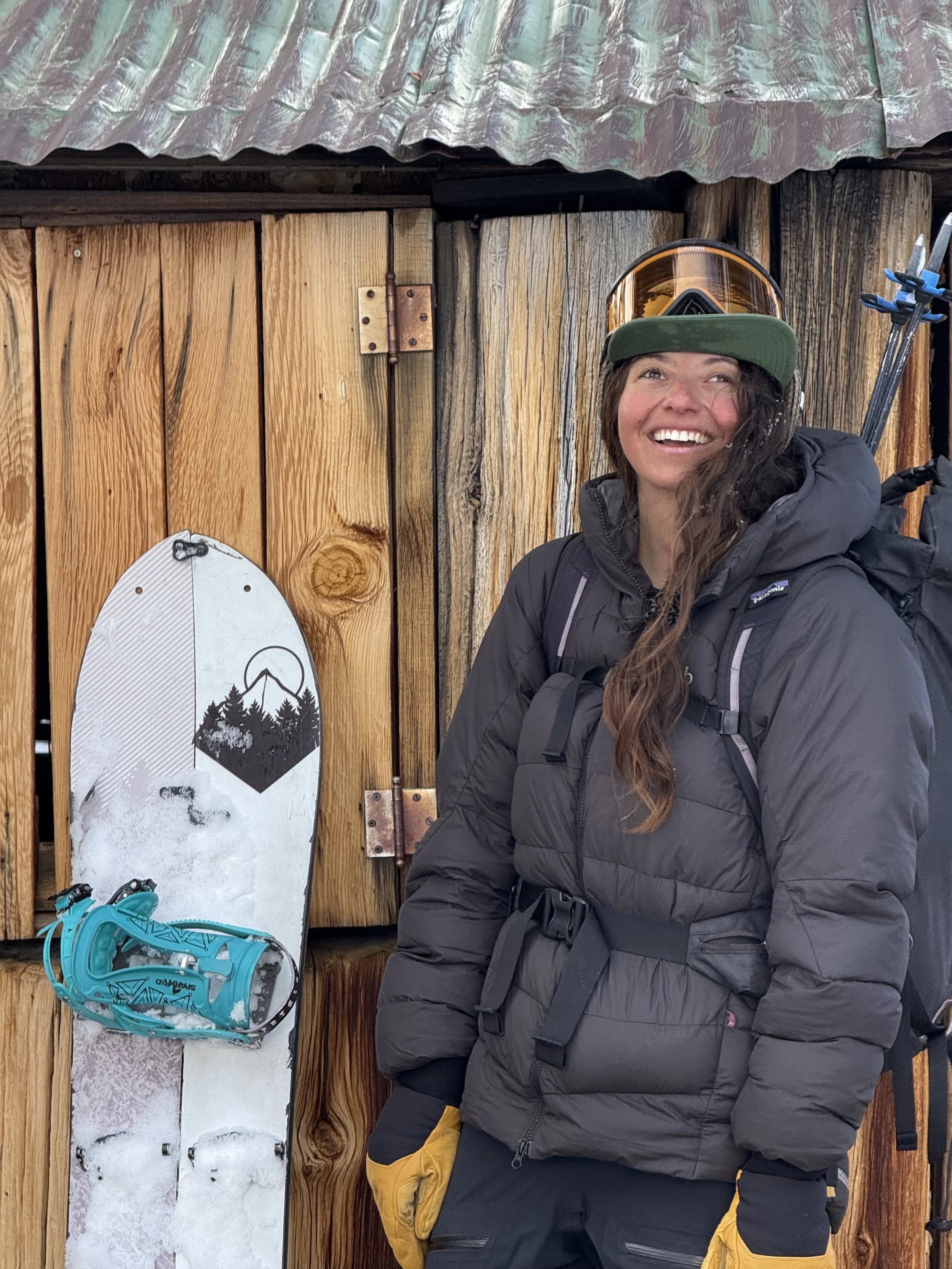 A smiling woman in winter gear, including a green cap, sunglasses, and a black jacket, standing in front of a wooden wall with a snowboard leaning against it.