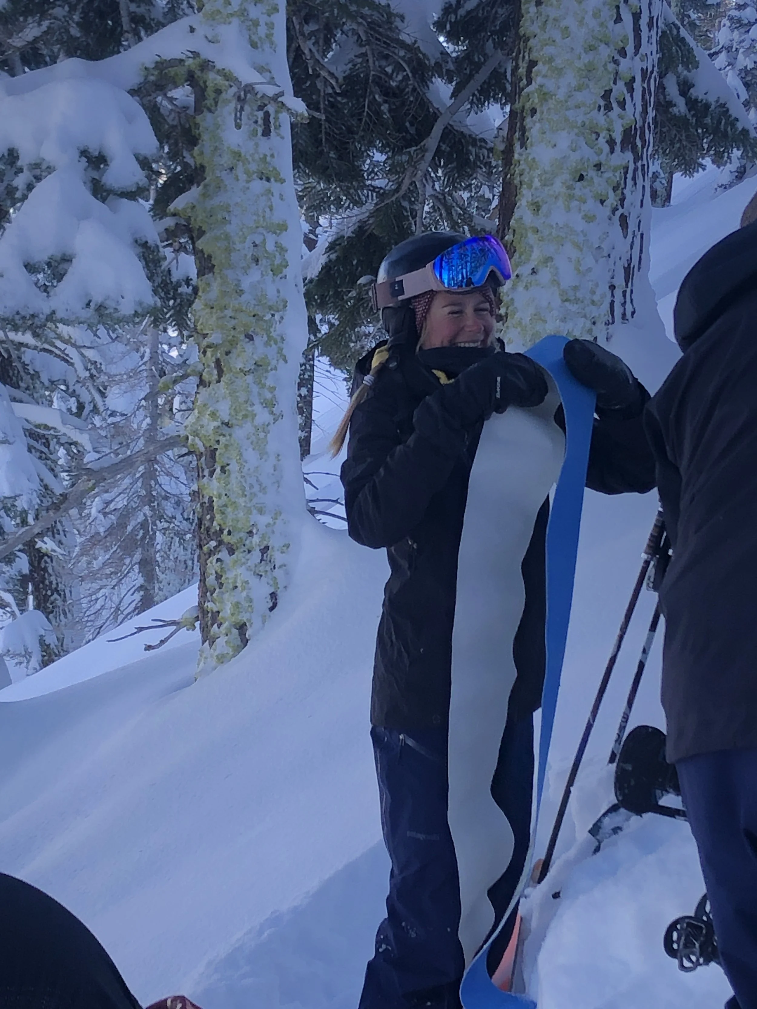 A woman wearing ski gear and goggles is laughing and holding a long piece of fabric or ribbon in a snowy, forested environment.