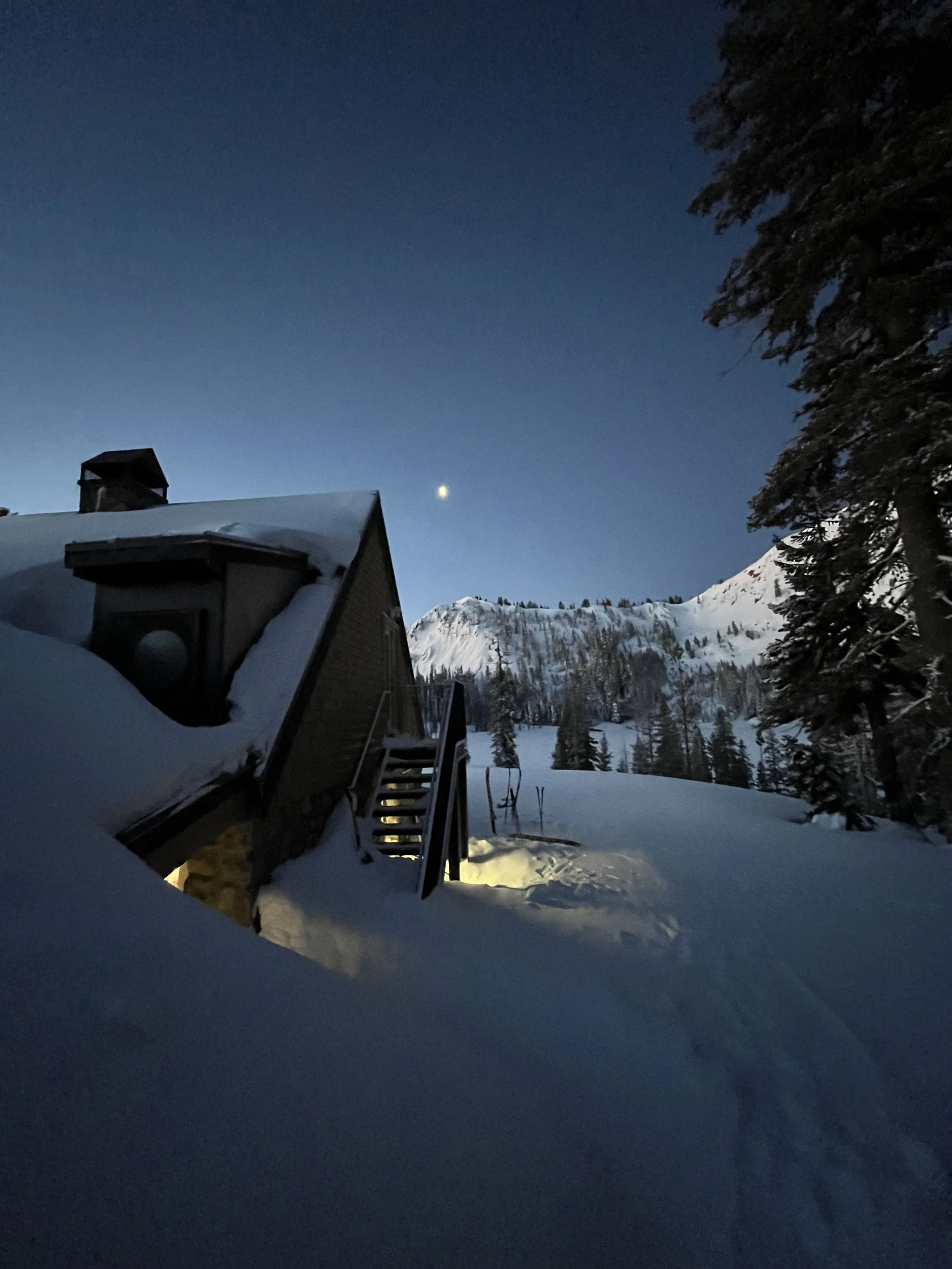 Snow-covered cabin at night with outdoor stairs, surrounded by winter landscape including snow and trees, under a dark blue sky with a bright celestial object.