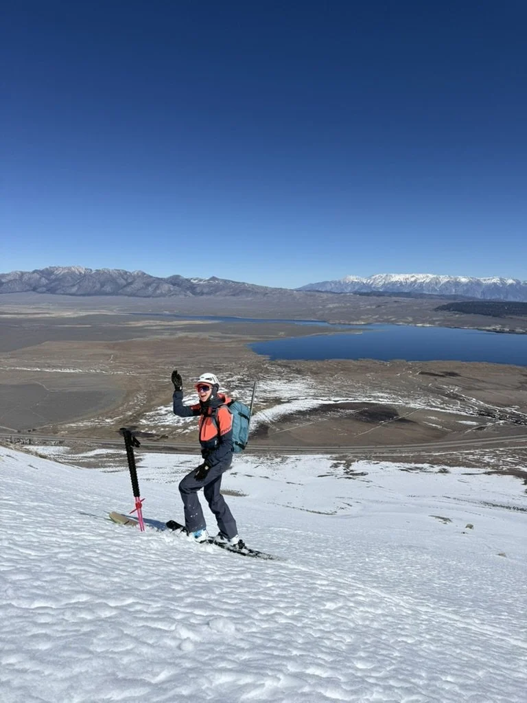 A person skiing up a snowy hill, waving, with mountains, a lake, and a clear blue sky in the background.