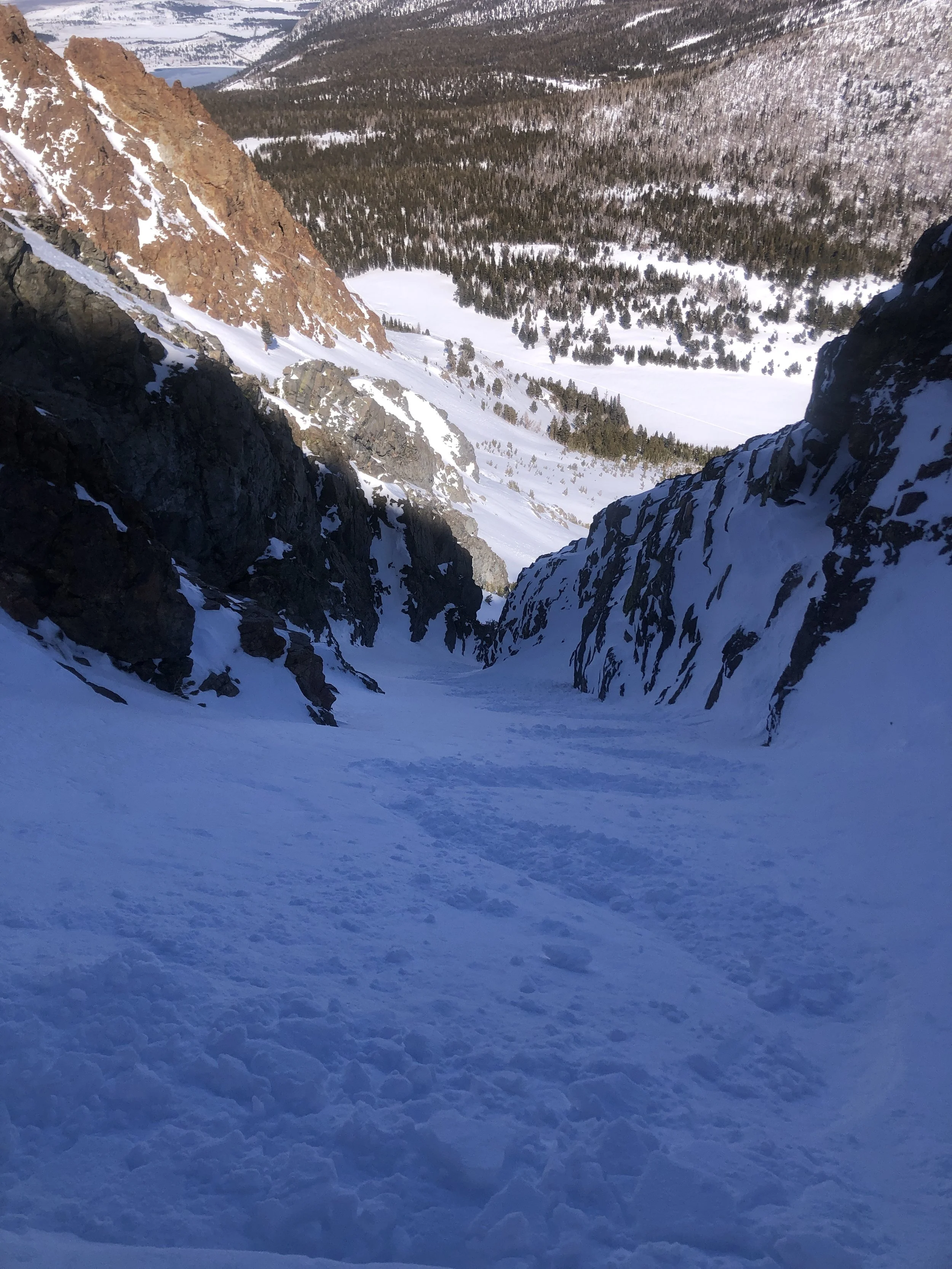 A snowy mountain landscape viewed from a high vantage point, with steep snow-covered slopes and rocky formations, leading down to a forested valley.