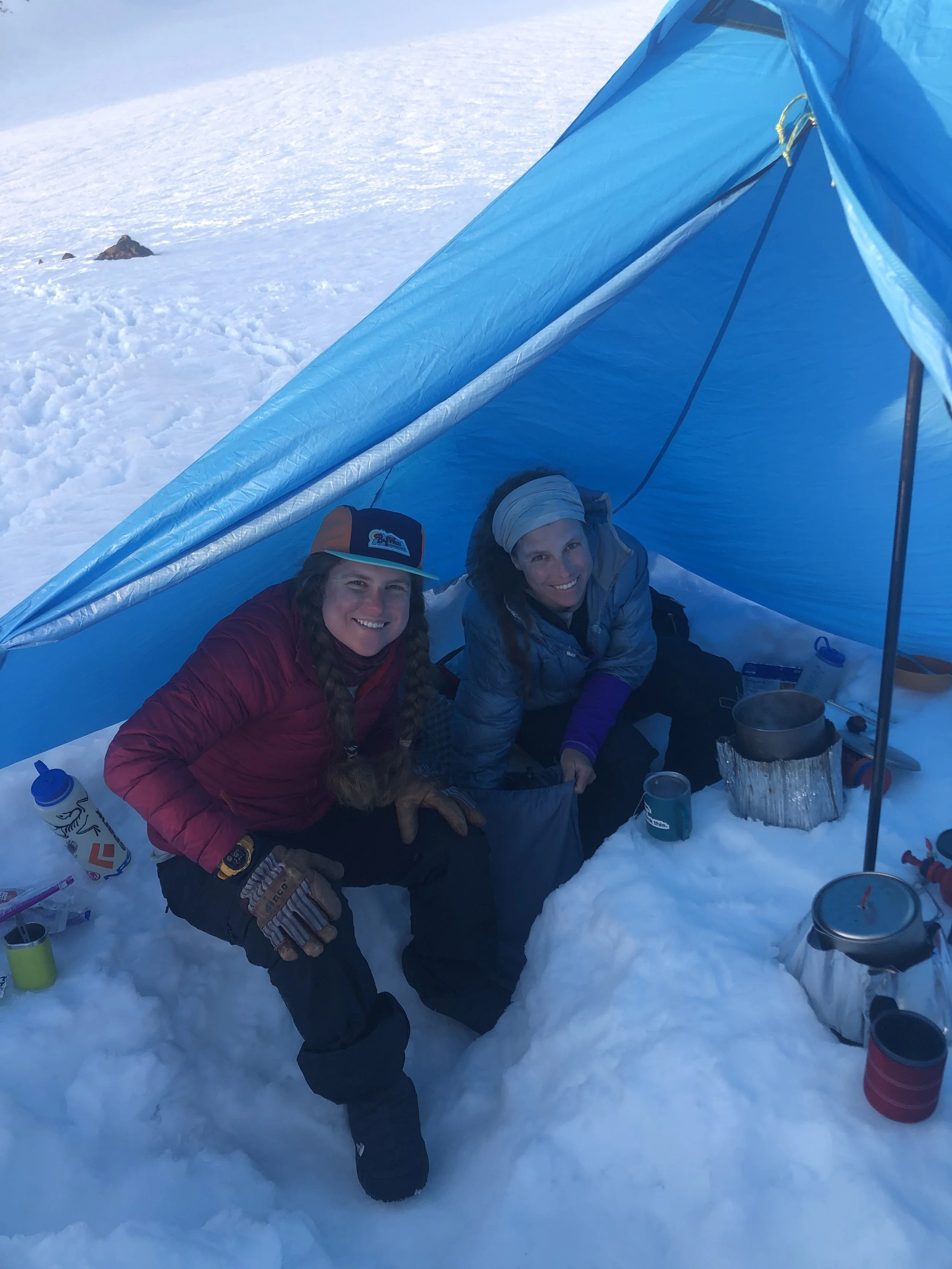 Two women sitting inside an ice shelter in a snowy landscape, smiling at the camera, with camping gear and supplies around them.