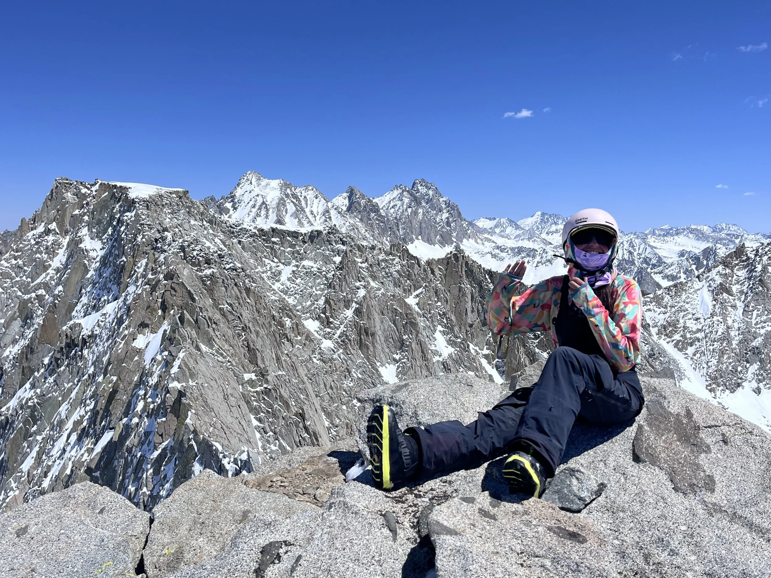 A person in colorful winter gear, wearing a helmet and goggles, sitting on a mountain ridge with snow-capped peaks and clear blue sky in the background.