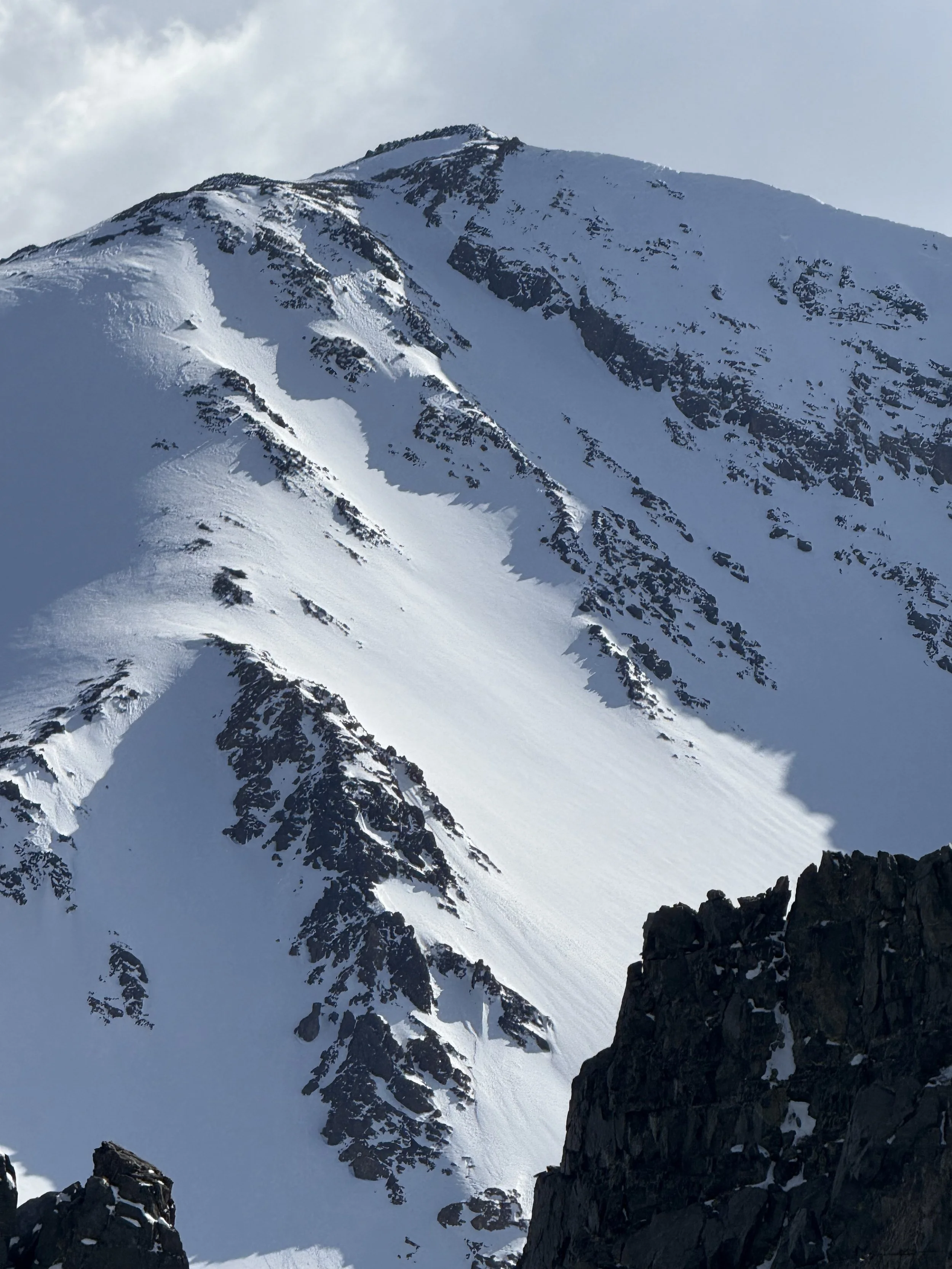 Snow-covered mountain with dark rock formations on the slopes and a partly cloudy sky.