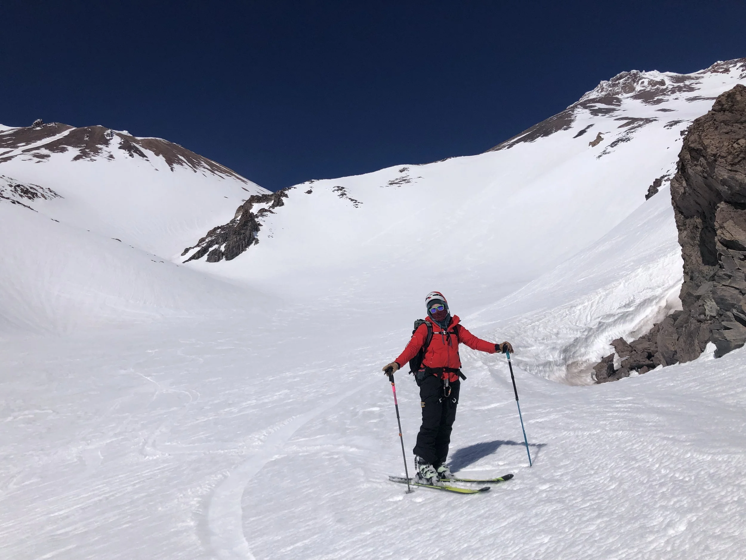 A person in red winter gear standing on skis in a snow-covered mountain landscape with a blue sky.