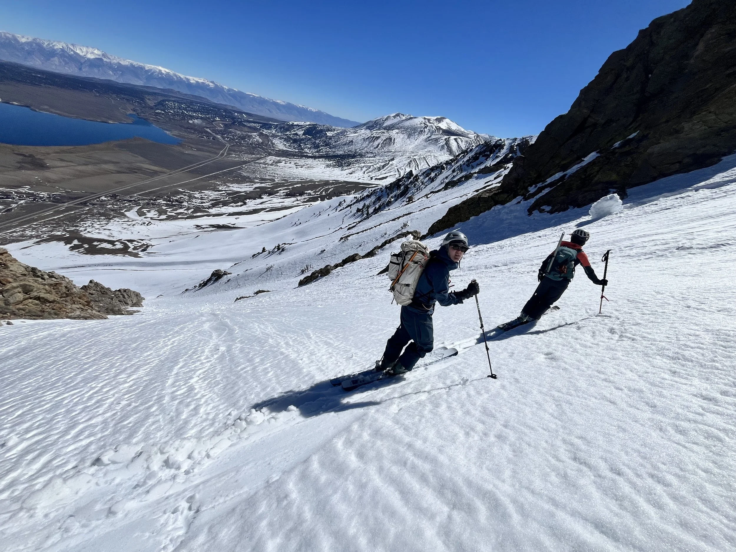 Two people skiing up a snowy mountain slope with a scenic view of mountains, a lake, and a clear blue sky in the background.
