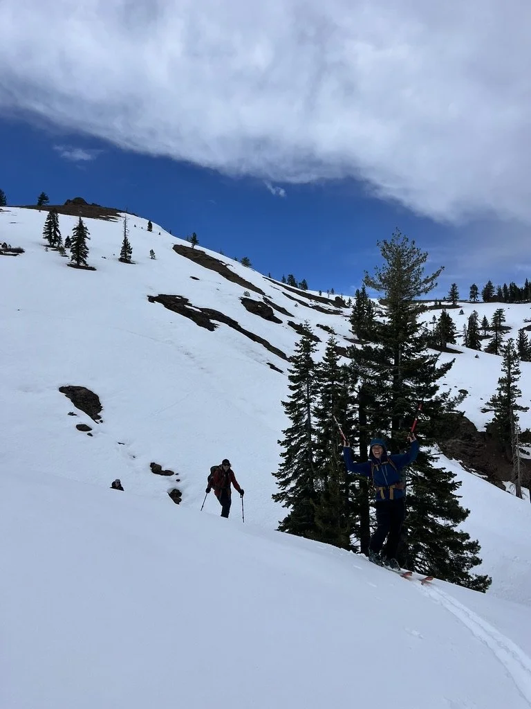 Two people skiing on a snow-covered mountain with trees and patches of rocks, under a partly cloudy sky.