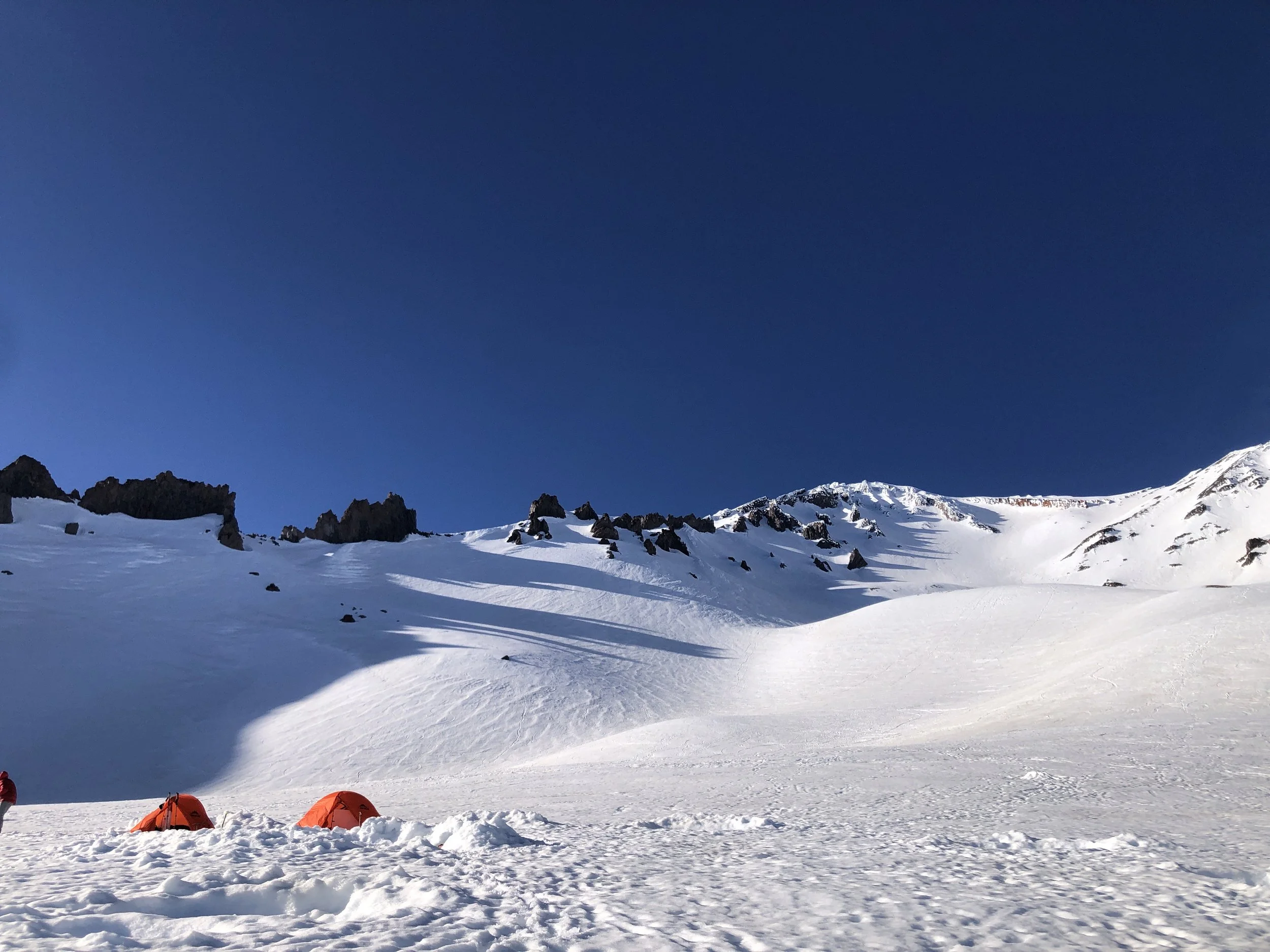 Two orange tents set up on snowy ground in a mountain landscape with snowy slopes, rocky outcroppings, and a clear blue sky.