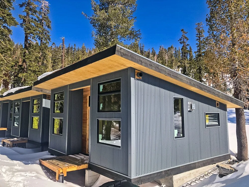 Modern tiny house with dark gray corrugated metal siding, multiple windows, wooden accents, and a flat roof, situated in a snowy forested area.