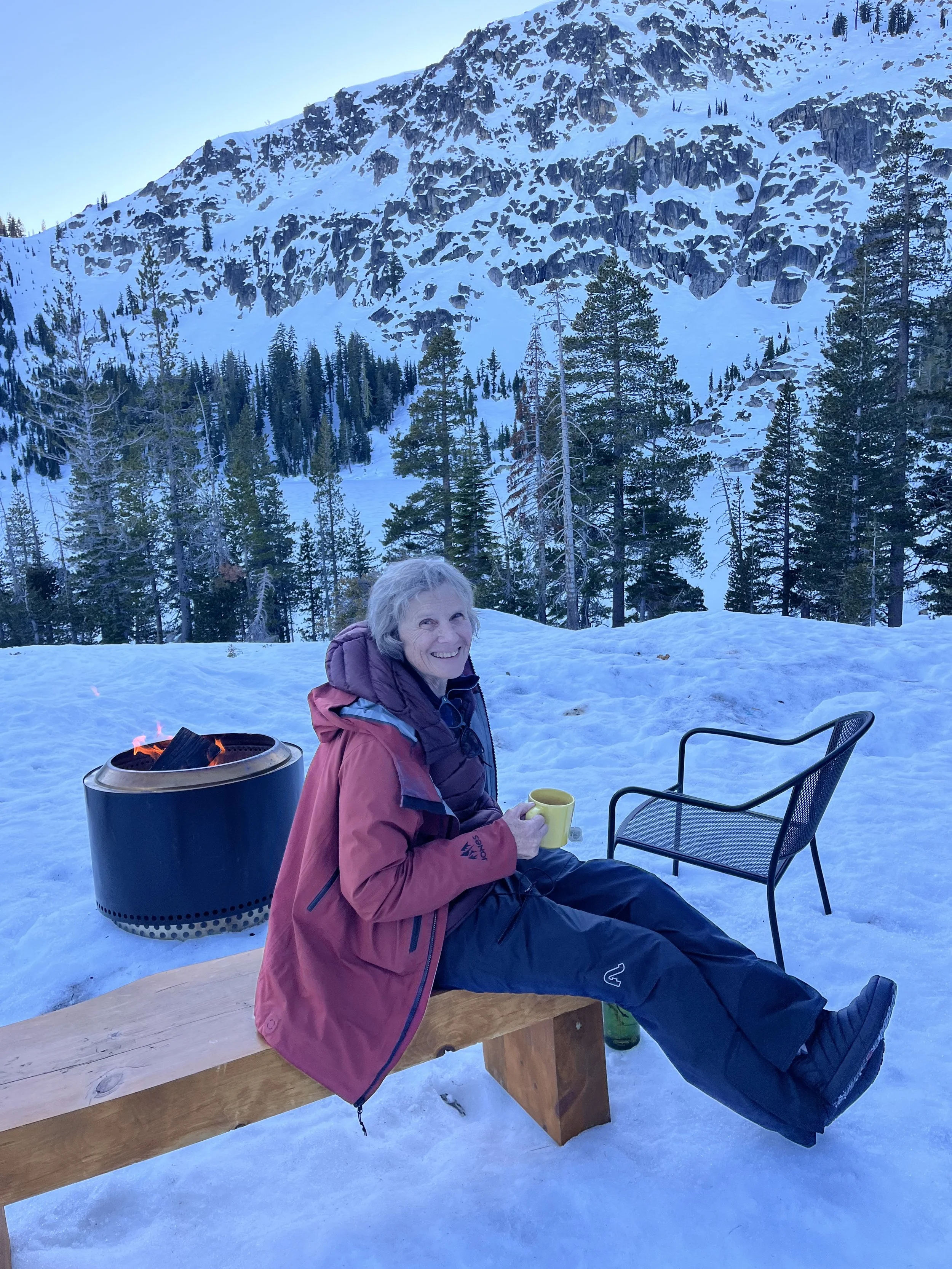 A smiling woman in a red winter jacket sitting on a wooden bench in a snowy outdoor setting with snow-covered trees and mountains in the background, holding a yellow mug.