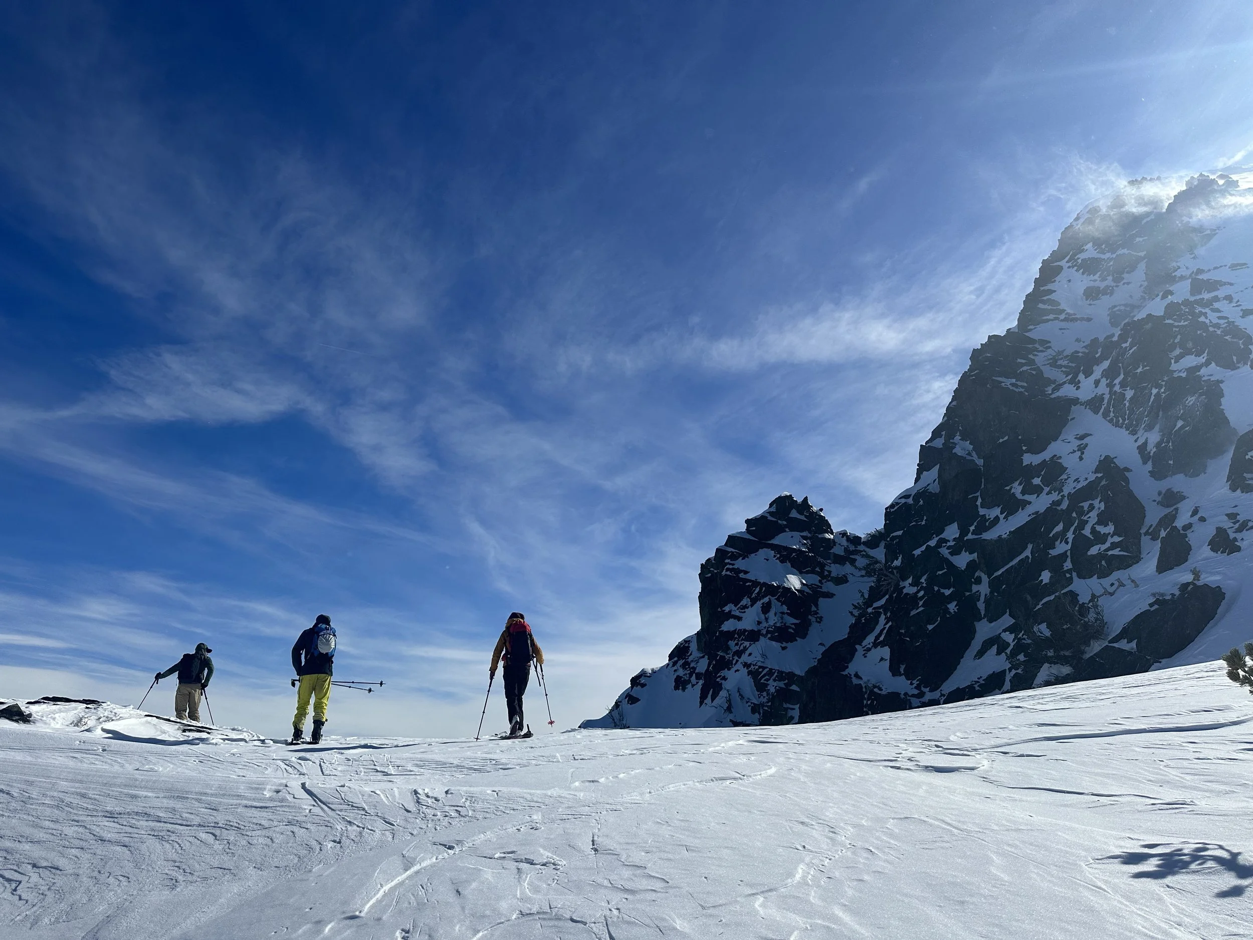 Three people mountaineering on snow-covered terrain with a prominent rocky, snow-covered mountain on the right and a blue sky with wispy clouds overhead.