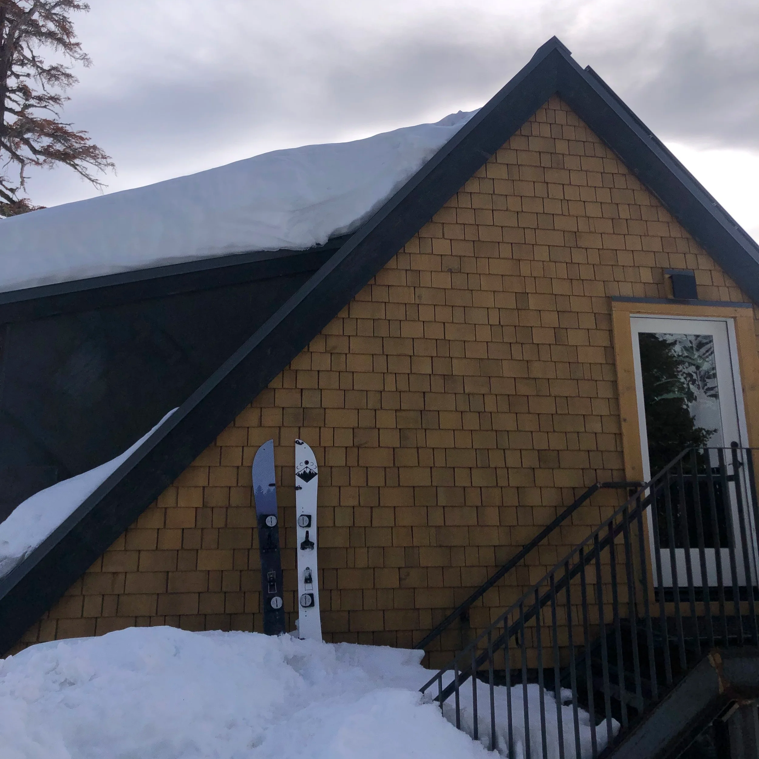 Snow-covered roof with a large snowdrift on top, a tree to the left, and a yellow-brick house with a window and stairs to the right, under a cloudy sky.