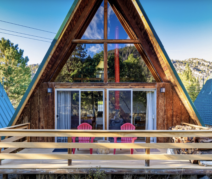Front view of an A-frame cabin with large triangular windows, a wooden balcony with two pink chairs, and a sliding glass door, surrounded by trees and a blue sky.