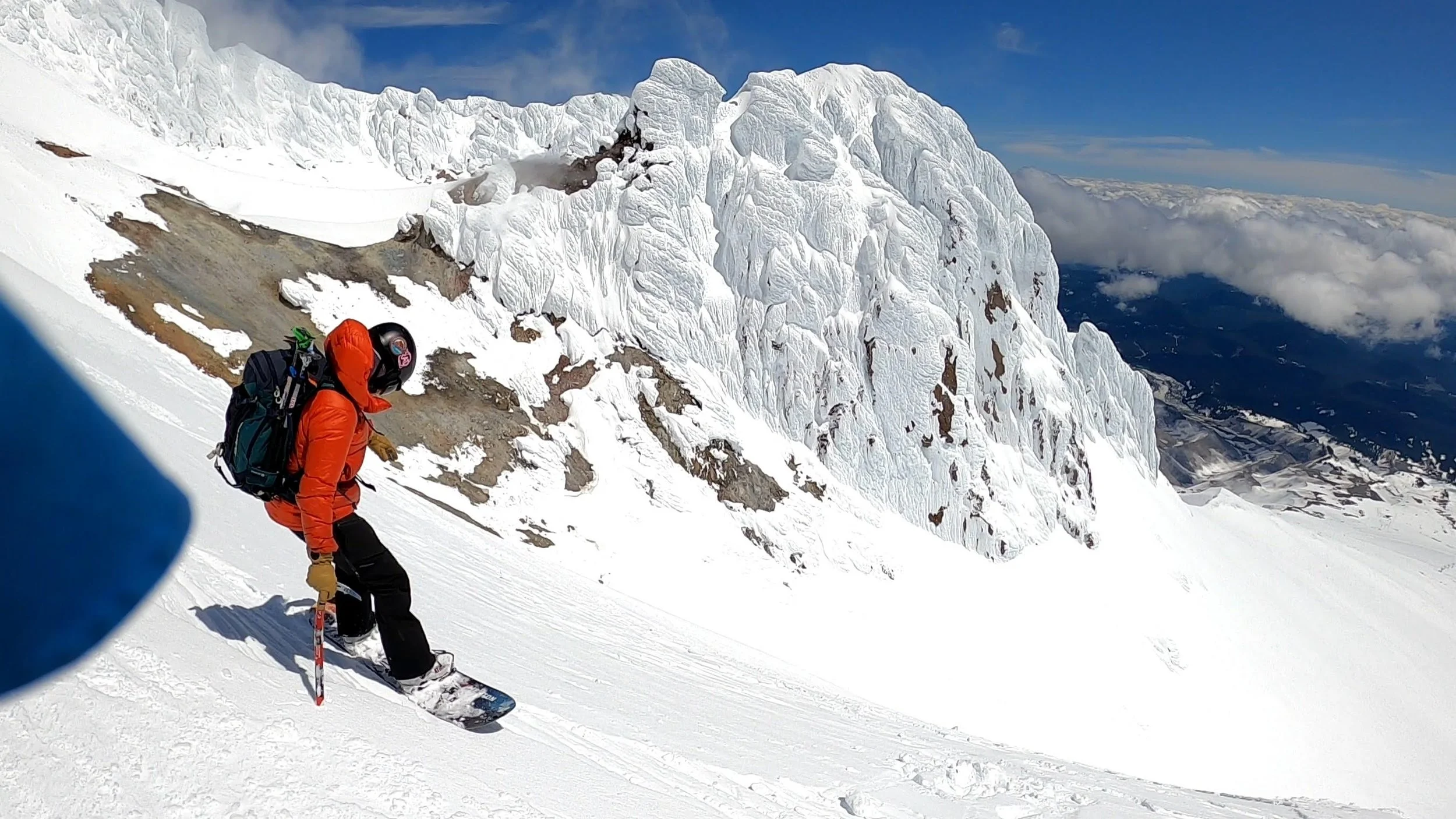 A person in an orange jacket and black pants snowboarding on a snowy mountain slope with a backpack, with a large icy mountain and clouds in the background.